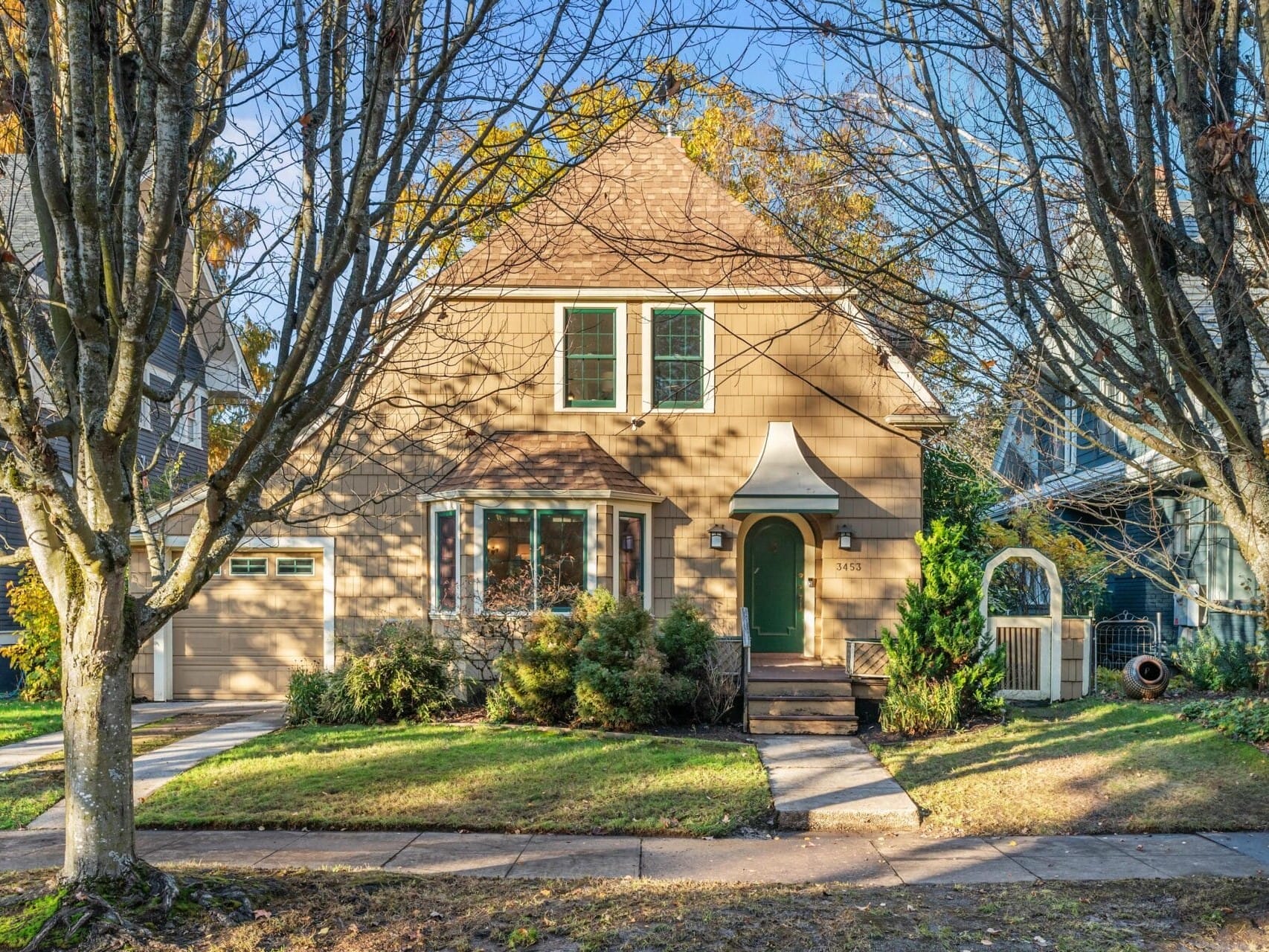 A charming two-story house with a pitched roof, set among bare trees. It features a brown exterior, a green door, and an attached garage. The front yard is grassy with a few bushes, and a clear blue sky is visible in the background.