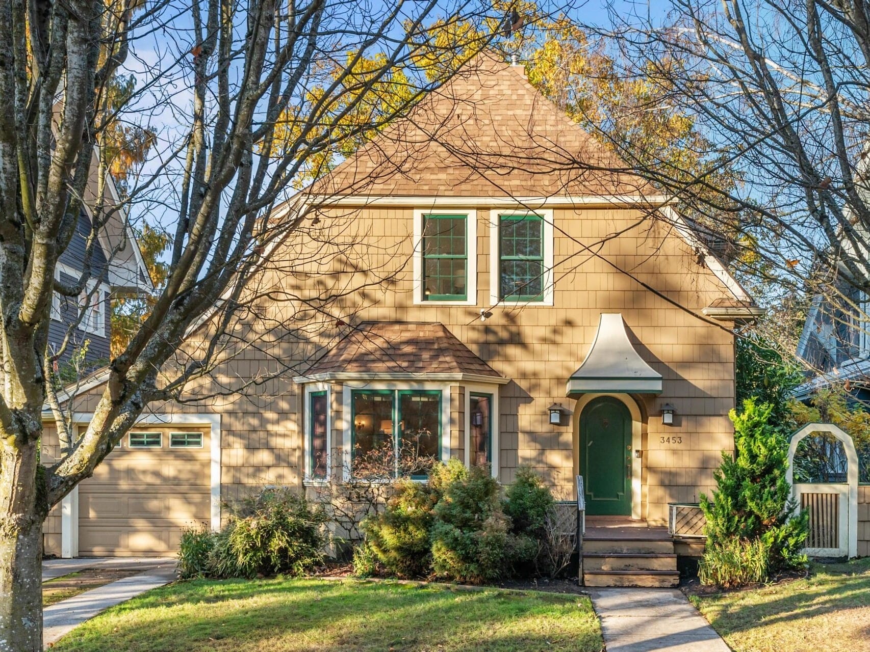 A charming two-story house with beige siding and a triangular roof, surrounded by trees. A path leads to the green front door, and theres a small porch canopy. A garage is attached on the left. The scene is bright and sunny.