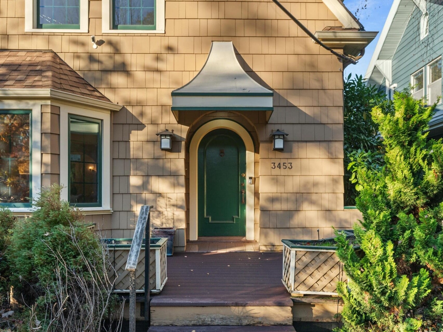 A cozy, light brown house with a green front door and two wall lamps. The house has a peaked awning and a small front porch surrounded by shrubs and a wooden railing. The street number 3453 is visible beside the door.