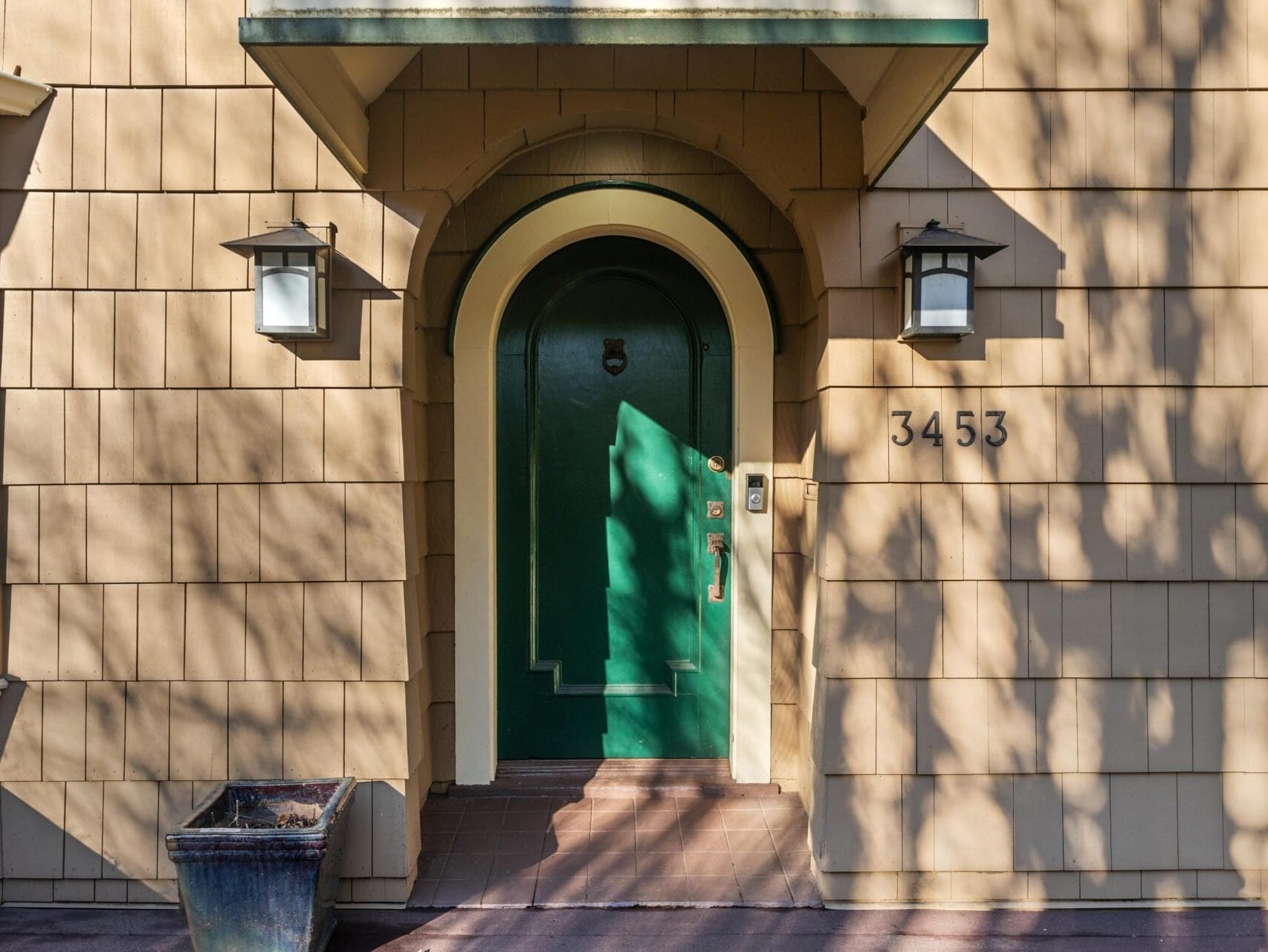 A tan house with a green arched door, flanked by two outdoor lanterns. The house number, 3453, is displayed to the right of the door. A planter sits to the left on the red-bricked entrance. Shadows of tree branches are cast on the house.