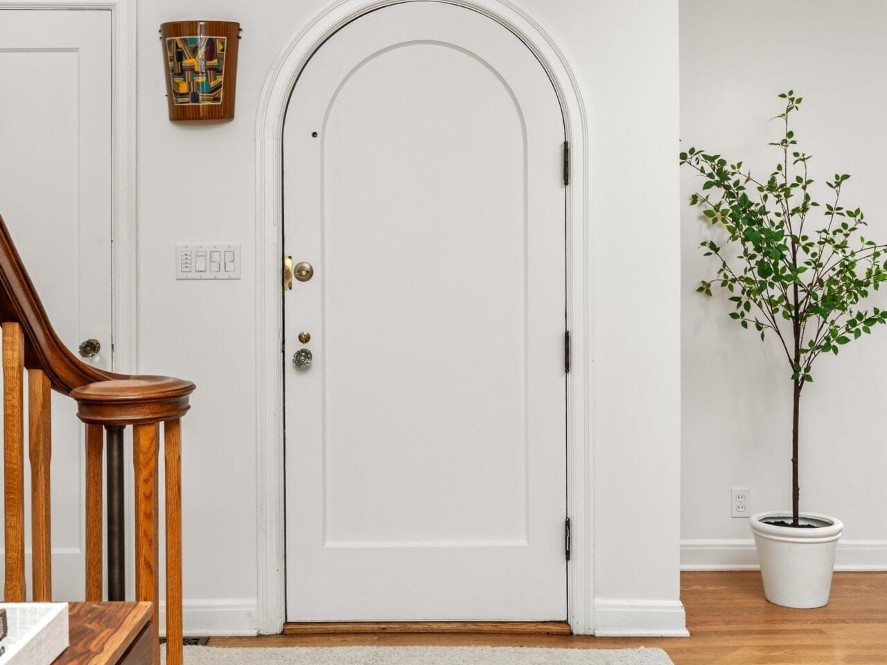 A white arched front door is centered in a room with light hardwood floors. A small potted plant stands to the right, while a wooden staircase with a smooth handrail is on the left. A colorful decorative plate hangs on the wall near the door.