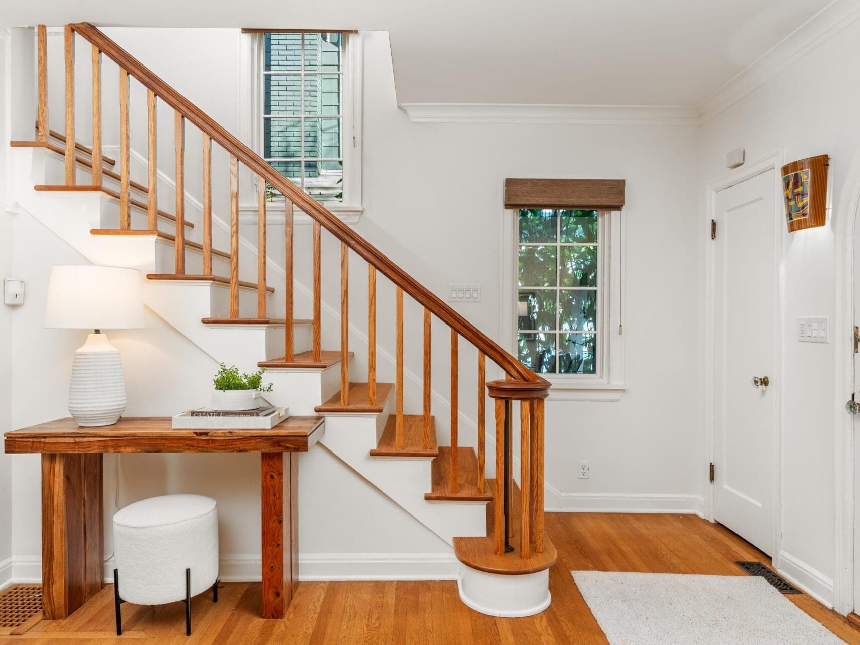 A bright foyer with wooden stairs and railing leading up. A wooden table with a lamp, plant, and book is beside the stairs. A small white stool is under the table. White walls, hardwood floors, and a window with a view of trees complete the scene.