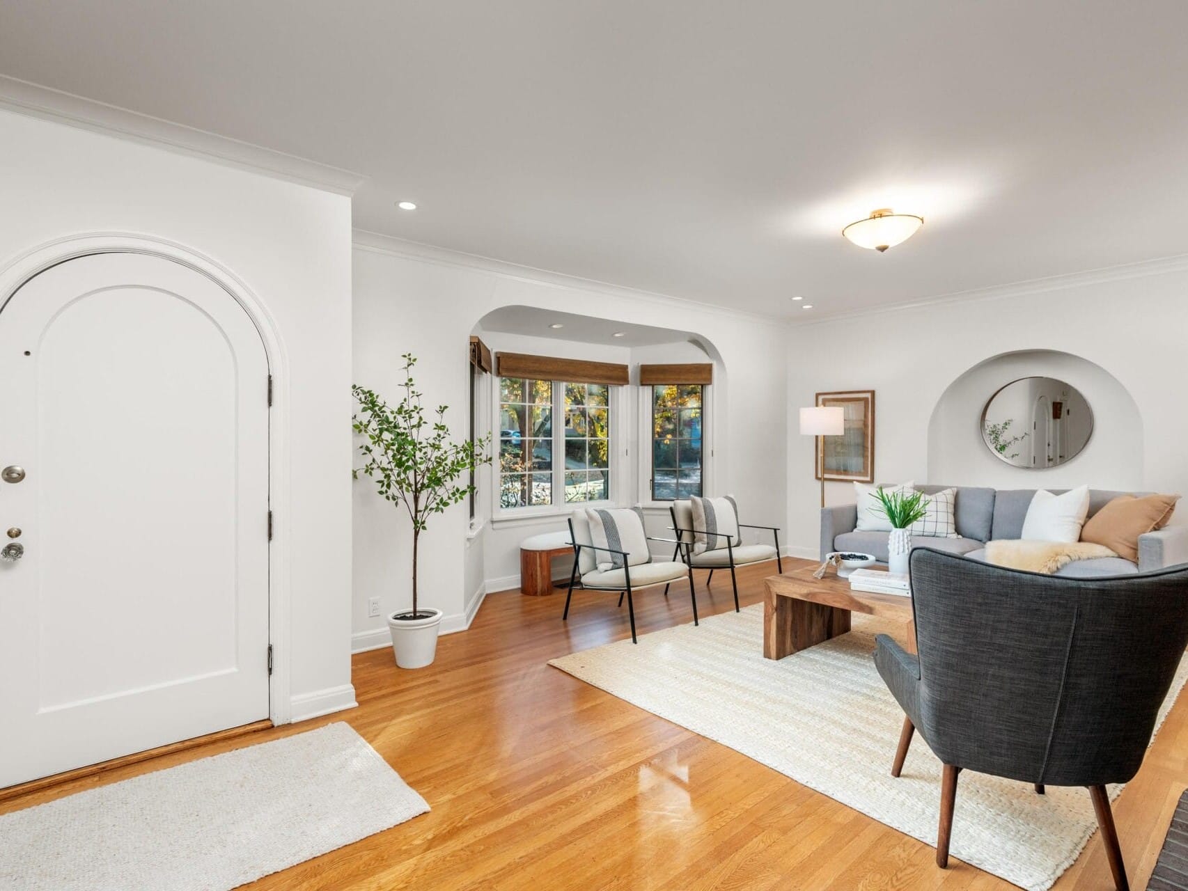 A modern living room with wooden floors, featuring a gray sofa, black armchairs, a wooden coffee table, and decorative plants. Natural light comes through a window with wooden blinds, and a round door is on the left. White walls and subtle lighting.