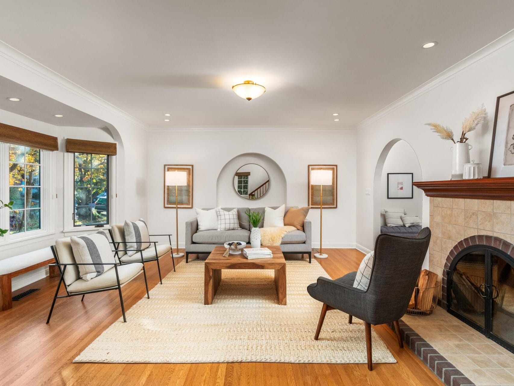 A cozy living room with a wooden floor, featuring a gray sofa, armchairs, and a wooden coffee table. A fireplace with a tiled surround is on the right, while large windows on the left bring in natural light. A round mirror and lamps enhance the space.