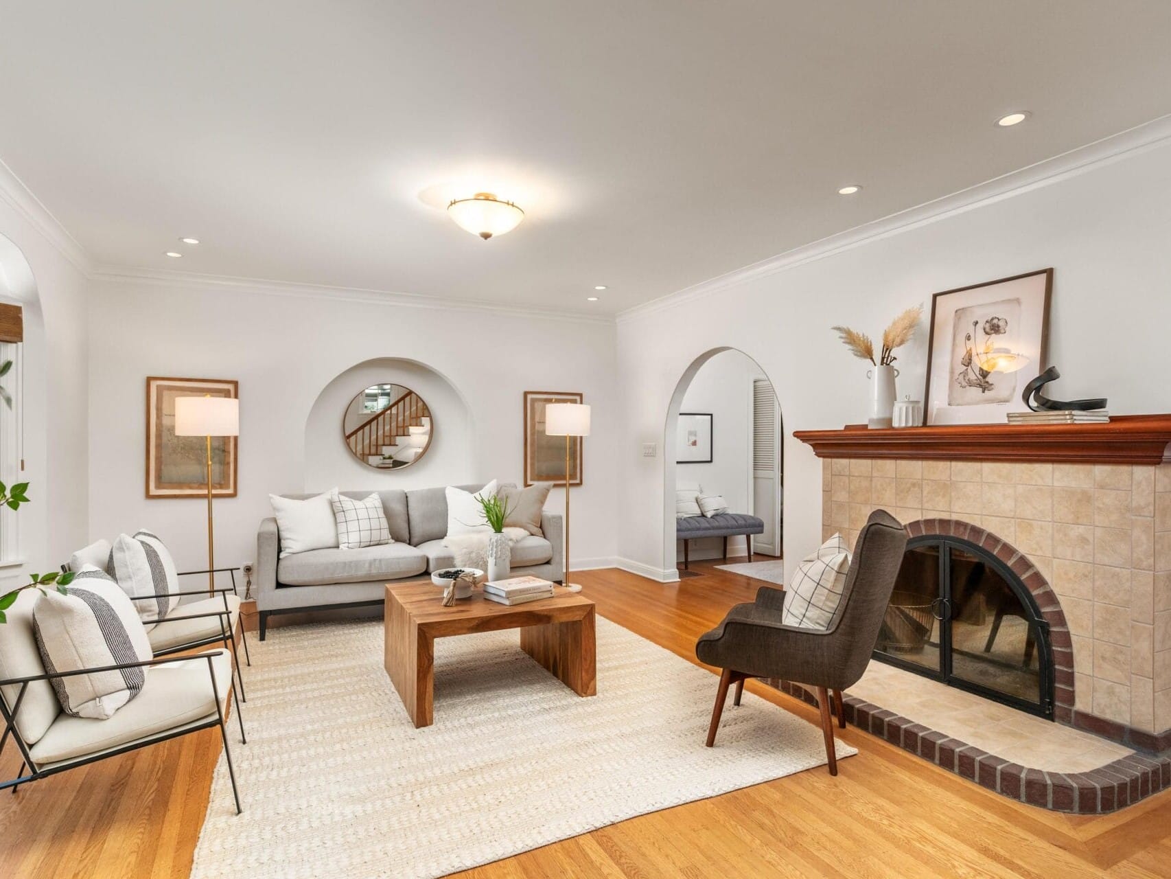 A cozy living room with a wooden floor features a gray sofa, two white armchairs, and a brown lounge chair. A fireplace with a wooden mantle is on the right, and a light rug covers the floor. Decorative mirrors and a framed artwork adorn the walls.