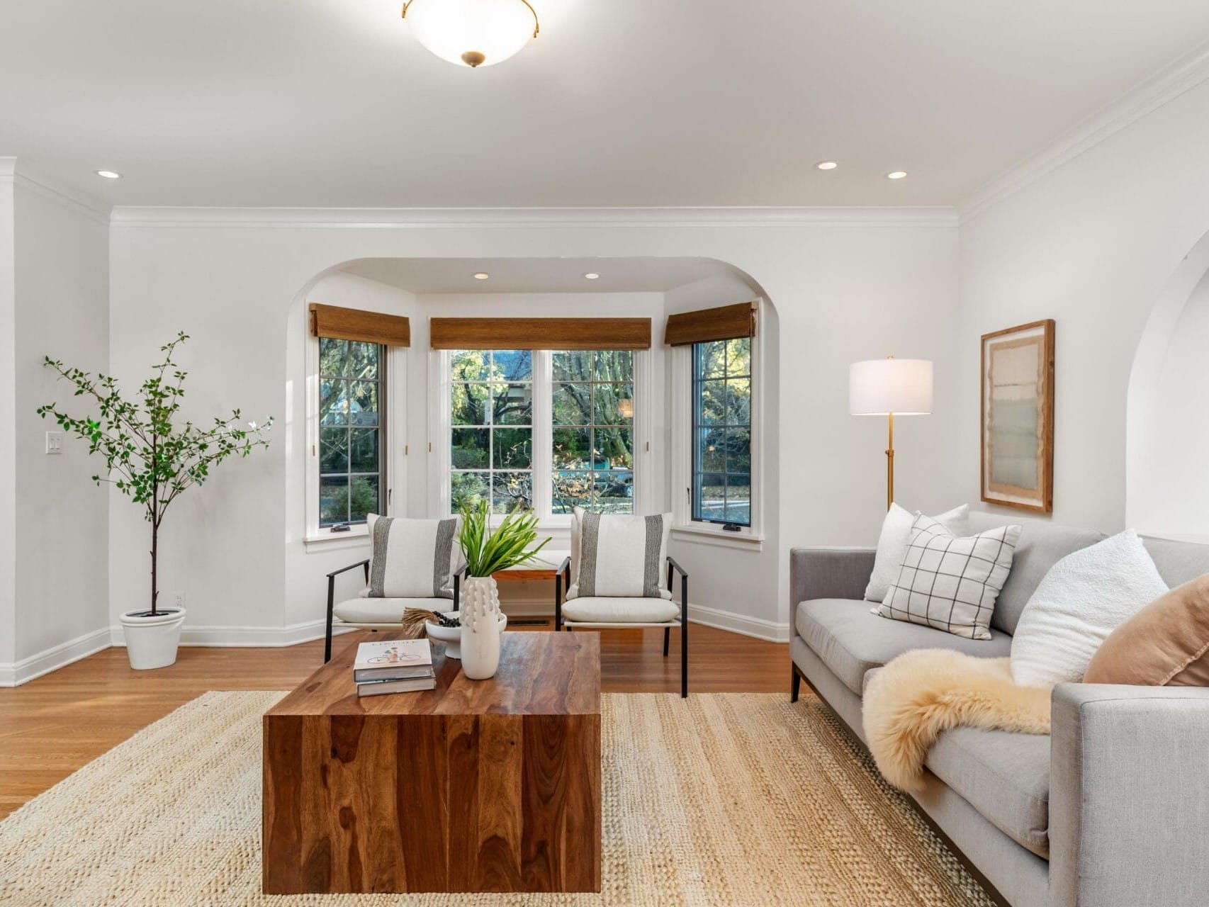 A cozy living room with a gray sofa, wooden coffee table, and patterned rug. Large windows with wooden blinds allow natural light. Decor includes a potted plant, floor lamp, and wall art, creating a welcoming atmosphere.