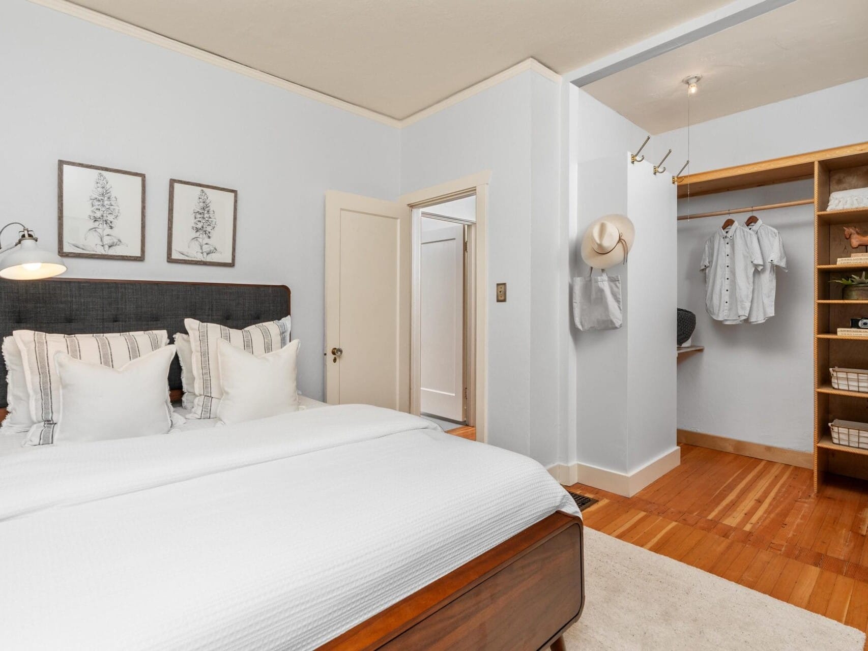 A neatly arranged bedroom with a wooden bed, white bedding, and decorative pillows. The wall features botanical prints, and theres an open closet with shirts and shelves. A hat and a shirt hang on the wall. The floor is hardwood.