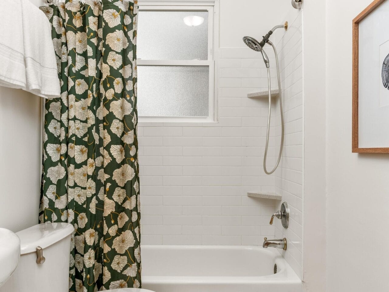 A small bathroom with a white bathtub and tiled shower, featuring a green shower curtain with a floral pattern. There is a white toilet and a towel on a rack. The floor appears to be tiled, and theres a framed picture on the wall.