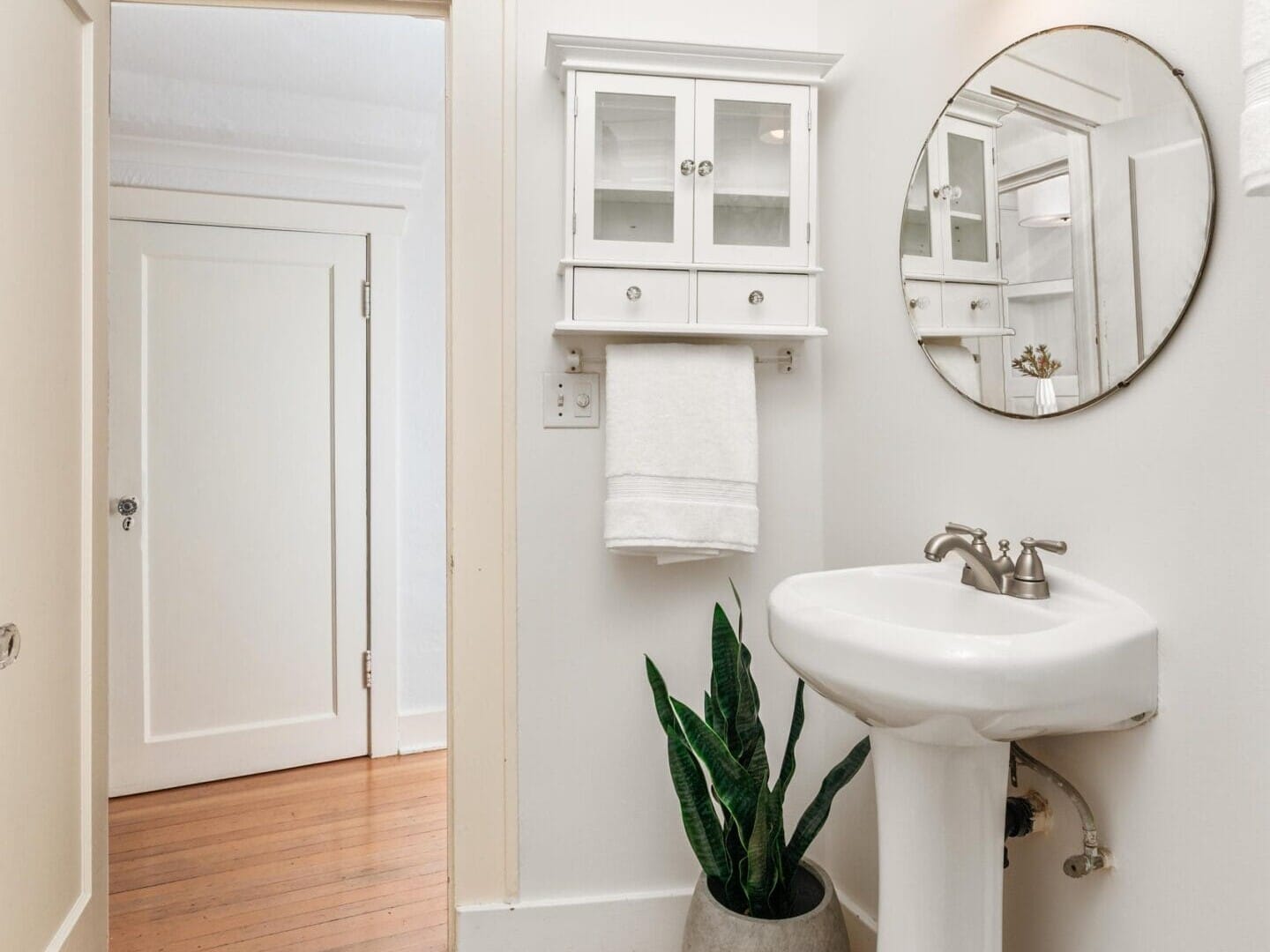 A small, minimalist bathroom with a white pedestal sink, round mirror, and wall-mounted cabinet. A potted plant sits on the floor next to the sink. A towel hangs beneath the cabinet, and the wooden door is open, revealing a hardwood floor.