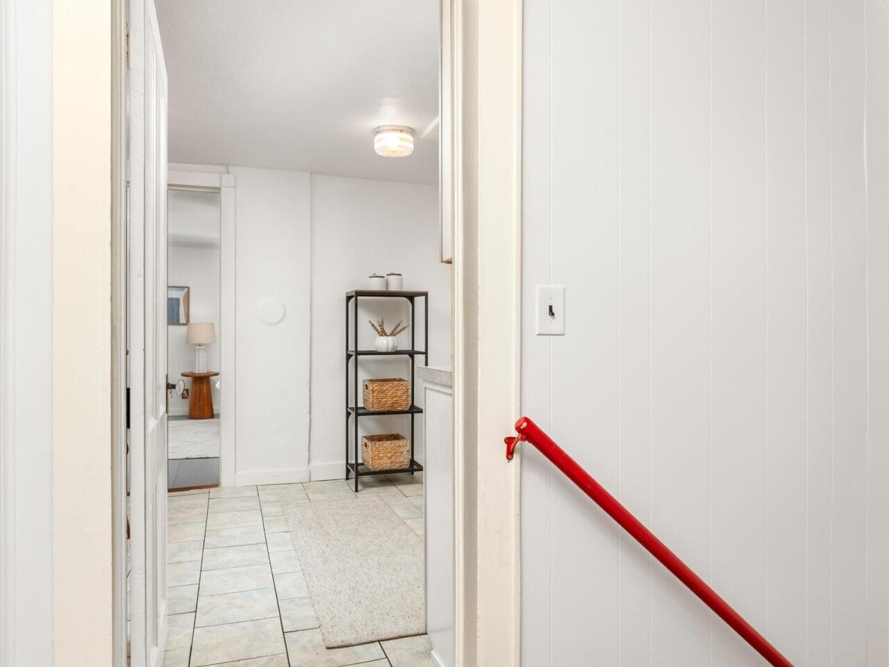 A staircase with a red handrail leads down to a room. The room has tiled flooring, a beige rug, and a metal shelf with wicker baskets. The walls are painted white and there is a light fixture on the ceiling.