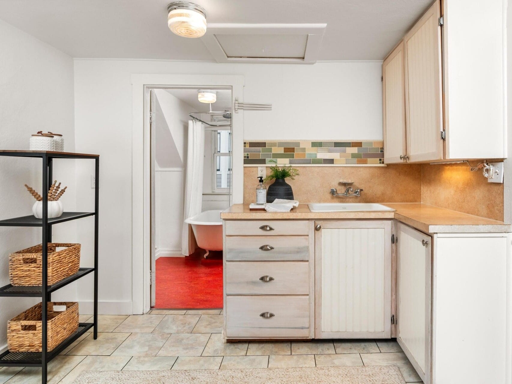 A small kitchen with white cabinets, wooden countertops, and a colorful tile backsplash. There is a black shelf with wicker baskets on the left and a visible adjoining room with a clawfoot bathtub on a red floor in the background.