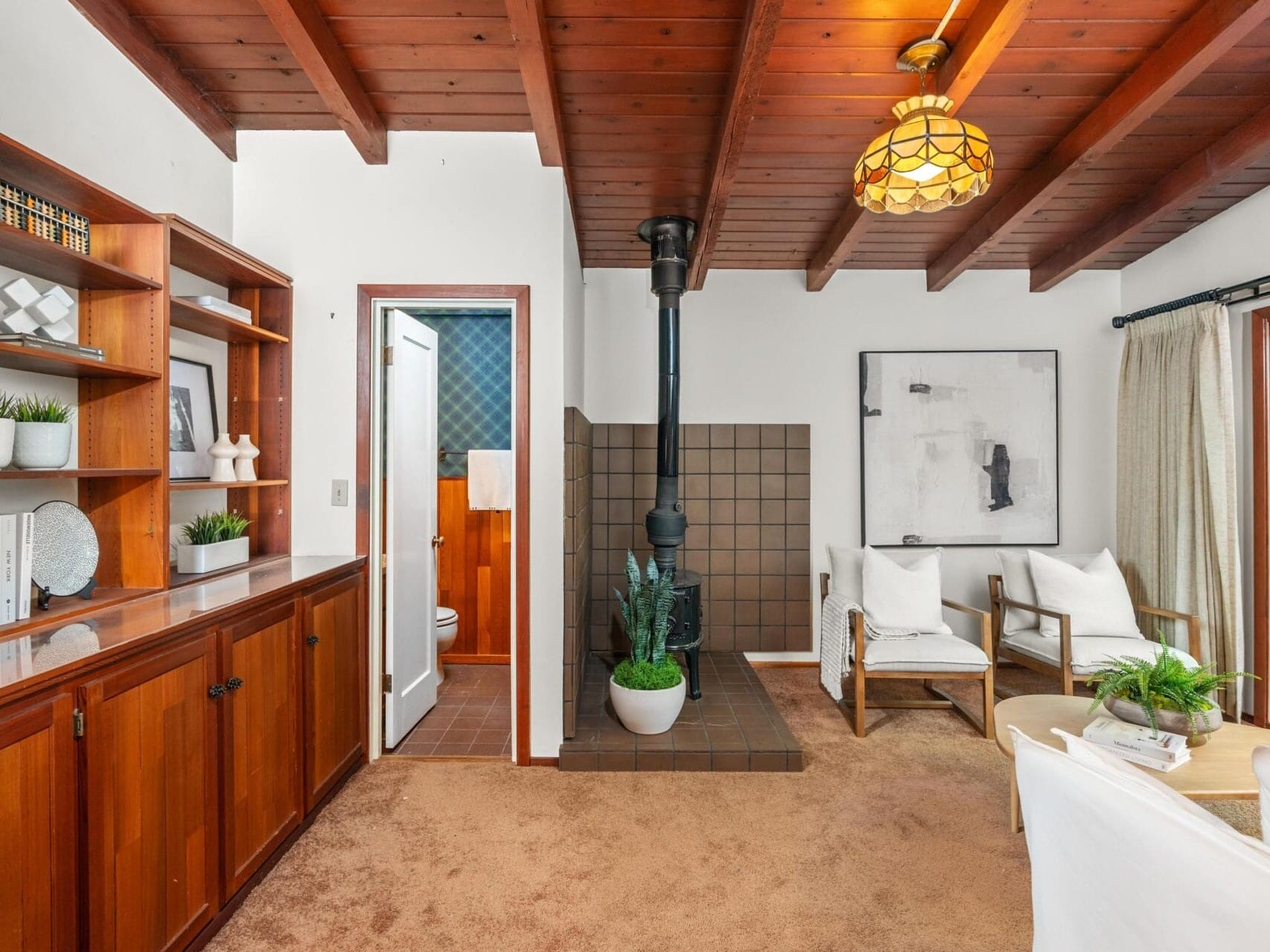 A cozy living room with wooden ceiling beams, a small fireplace, and built-in wooden shelves. There are white chairs, a modern artwork on the wall, and a large window with curtains. A door leads to a small, partially visible bathroom.