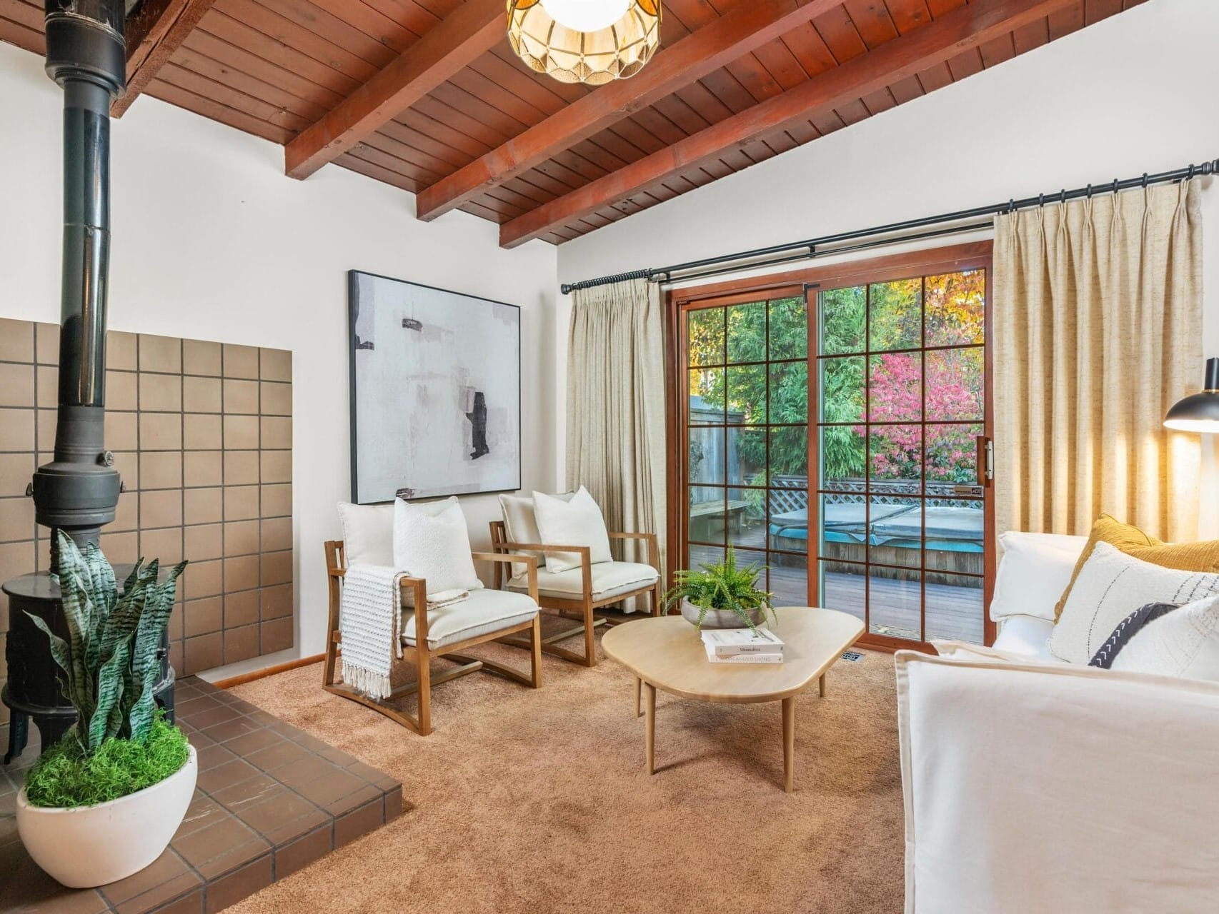 Cozy living room with a wood-burning stove, white chairs, and a sofa, centered around a wooden coffee table. Large glass doors lead to a deck and garden outside. Exposed wooden beams and a potted plant add to the rustic charm.