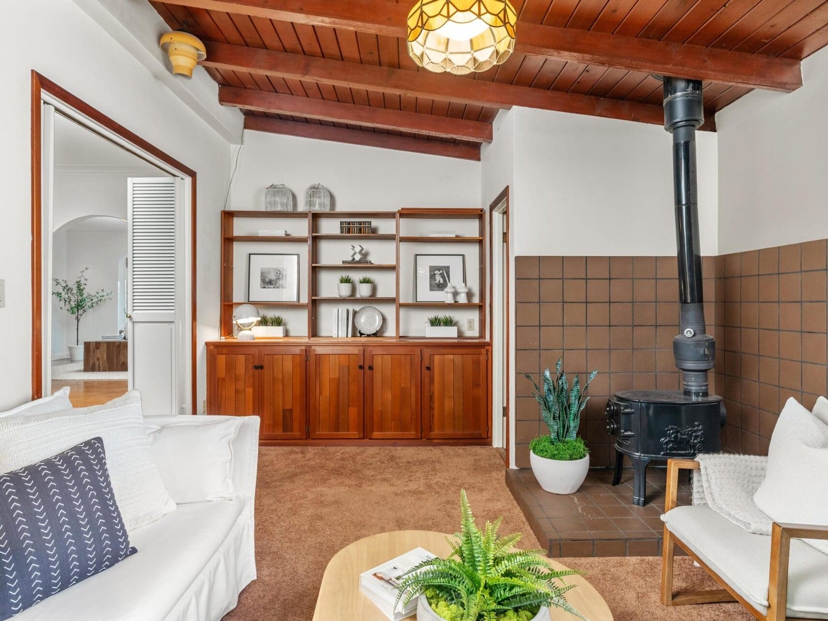 A cozy living room with a white sofa, wooden ceiling, and a wood-burning stove. Theres a wooden cabinet with shelves holding plants and framed photos. A small table with a plant sits on the carpeted floor. A doorway leads to another room.