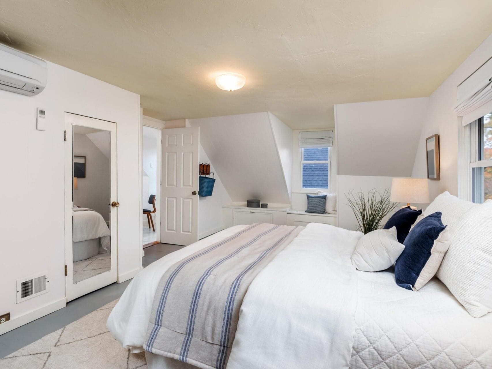 A cozy bedroom with a large bed covered in white and striped bedding. The room features white walls, a small window seat, and a wall-mounted air conditioner. A door leads to another room, and soft lighting enhances the serene atmosphere.