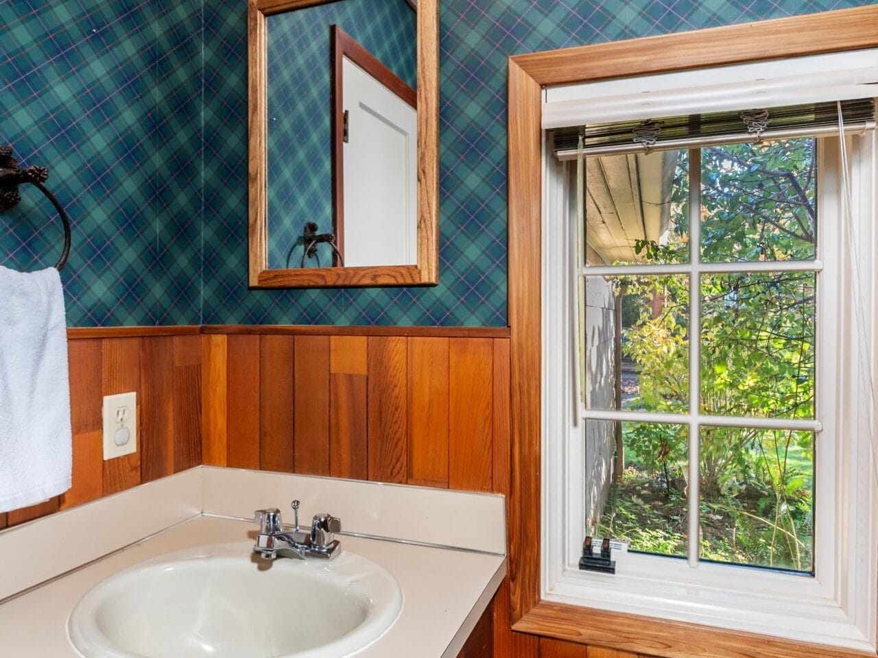 A small bathroom with a wood-paneled vanity and light-colored countertop. Above is a rectangular mirror. The walls have blue and green plaid wallpaper. A window with a view of greenery is beside the toilet.