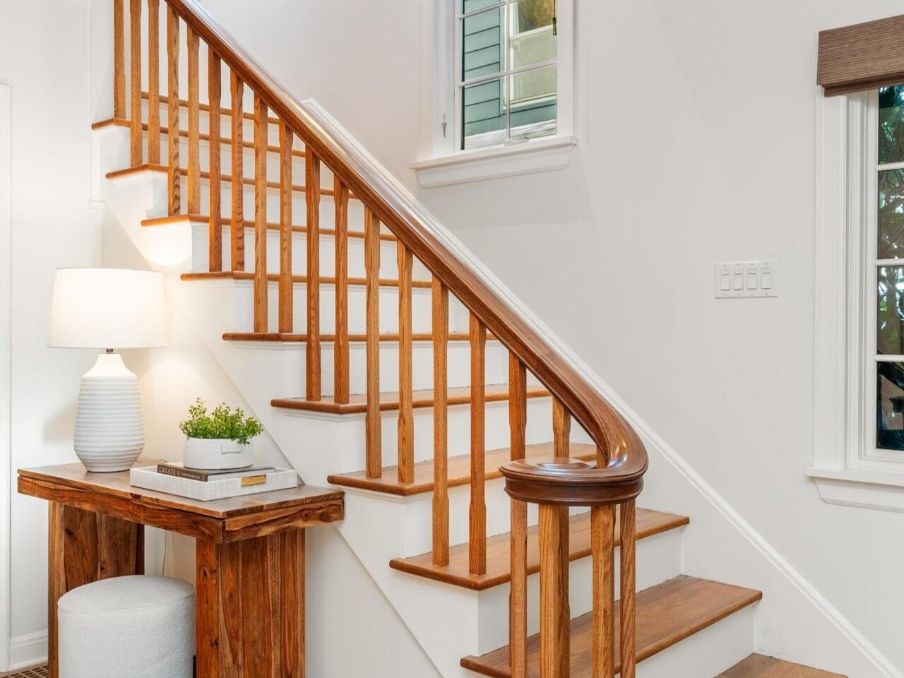 A wooden staircase with a polished handrail leads to the upper floor. A small wooden desk with a white lamp and a plant is positioned beneath the stairs. The room has white walls and a window with horizontal slats.