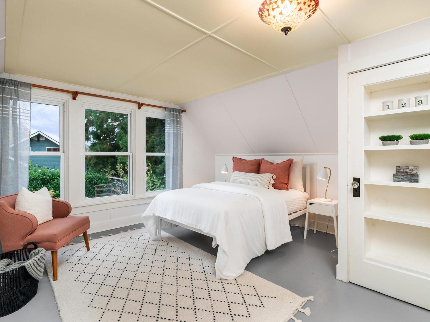 A cozy bedroom with a white bed, pink cushions, and a white nightstand. A pink armchair sits beside a window with sheer curtains. A patterned rug covers the gray floor, and a built-in shelf holds small decor items. A hanging light fixture illuminates the room.