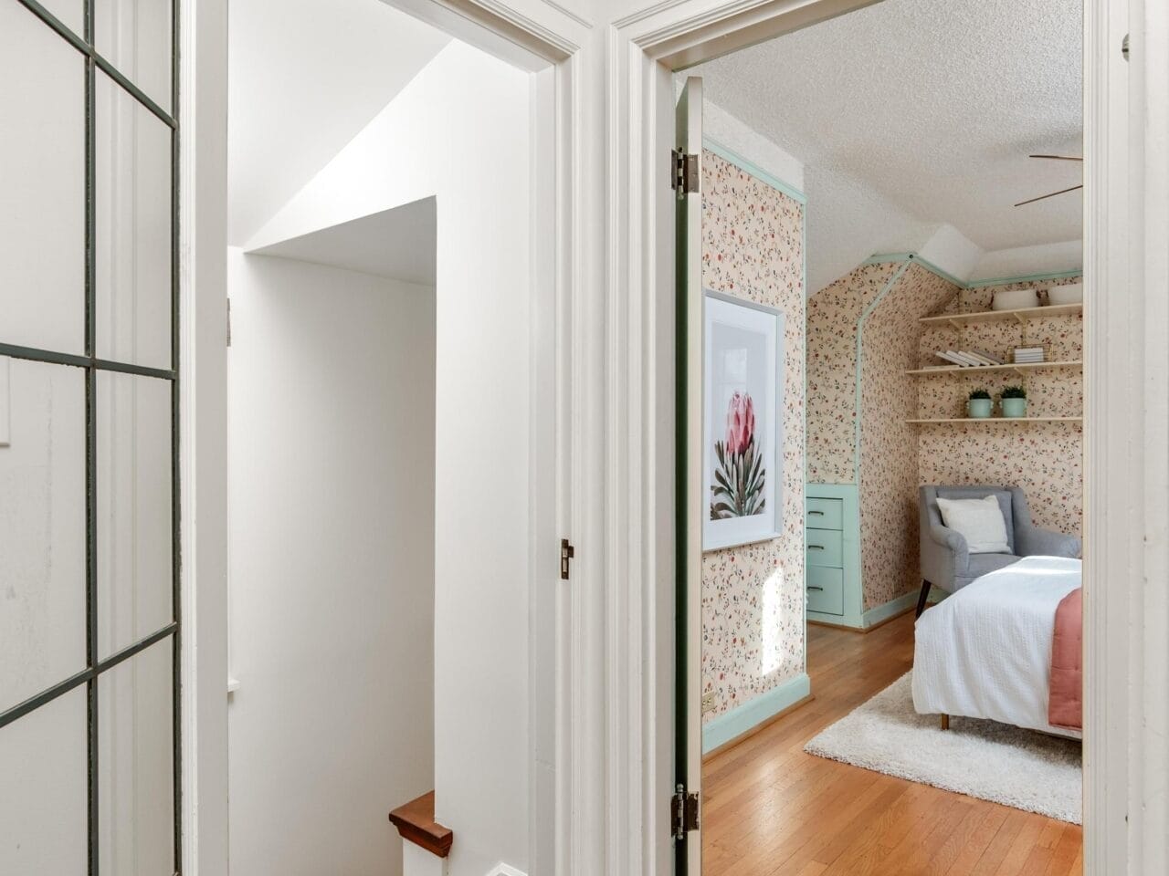 A hallway with wooden floors leads to a bedroom and a staircase. The bedroom has flower-patterned wallpaper, a neatly made bed, and built-in shelves. The door on the left leads down a flight of stairs.