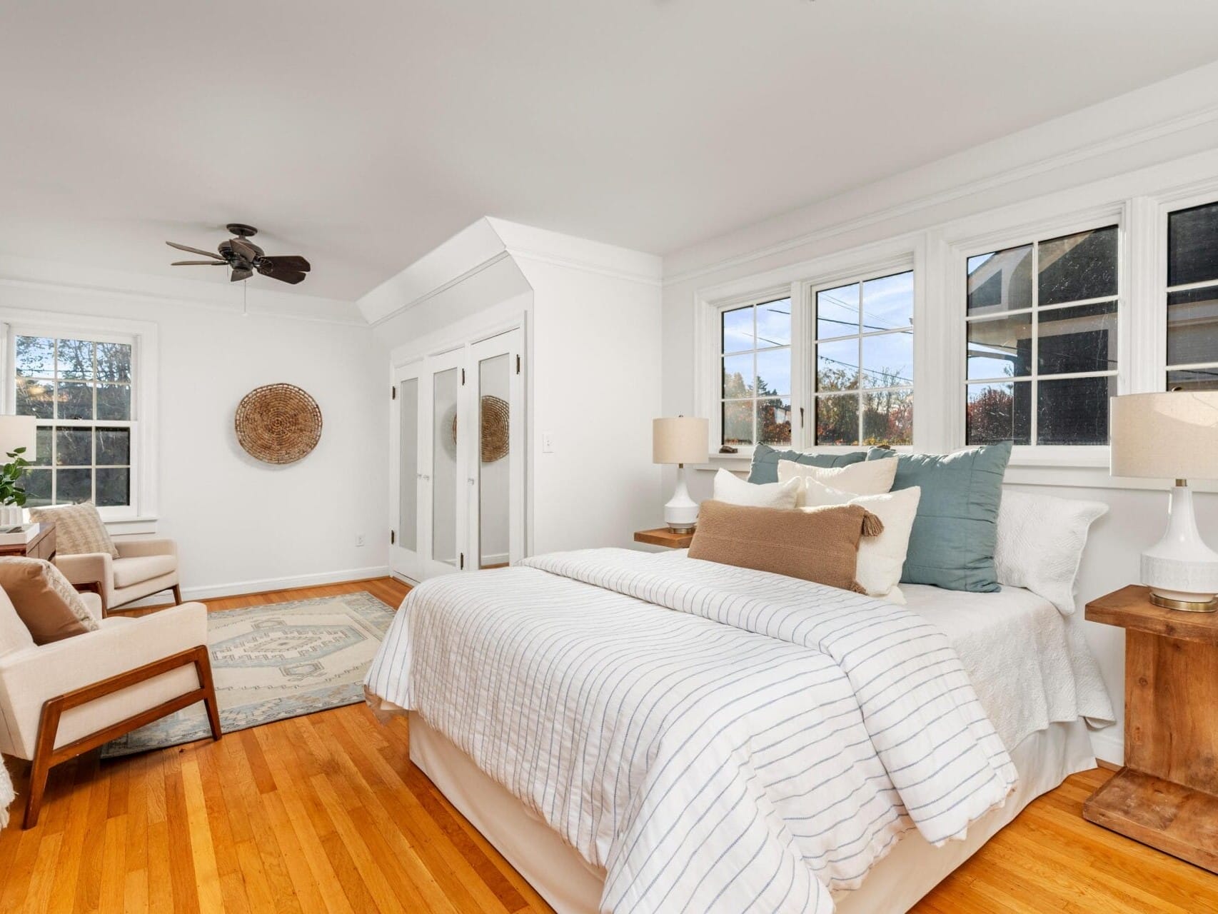 A bright, airy bedroom with a neatly made bed adorned with pillows. Two cozy chairs sit by a window. The room features a ceiling fan, wooden floor, and a woven wall decoration. Natural light fills the space through large windows.