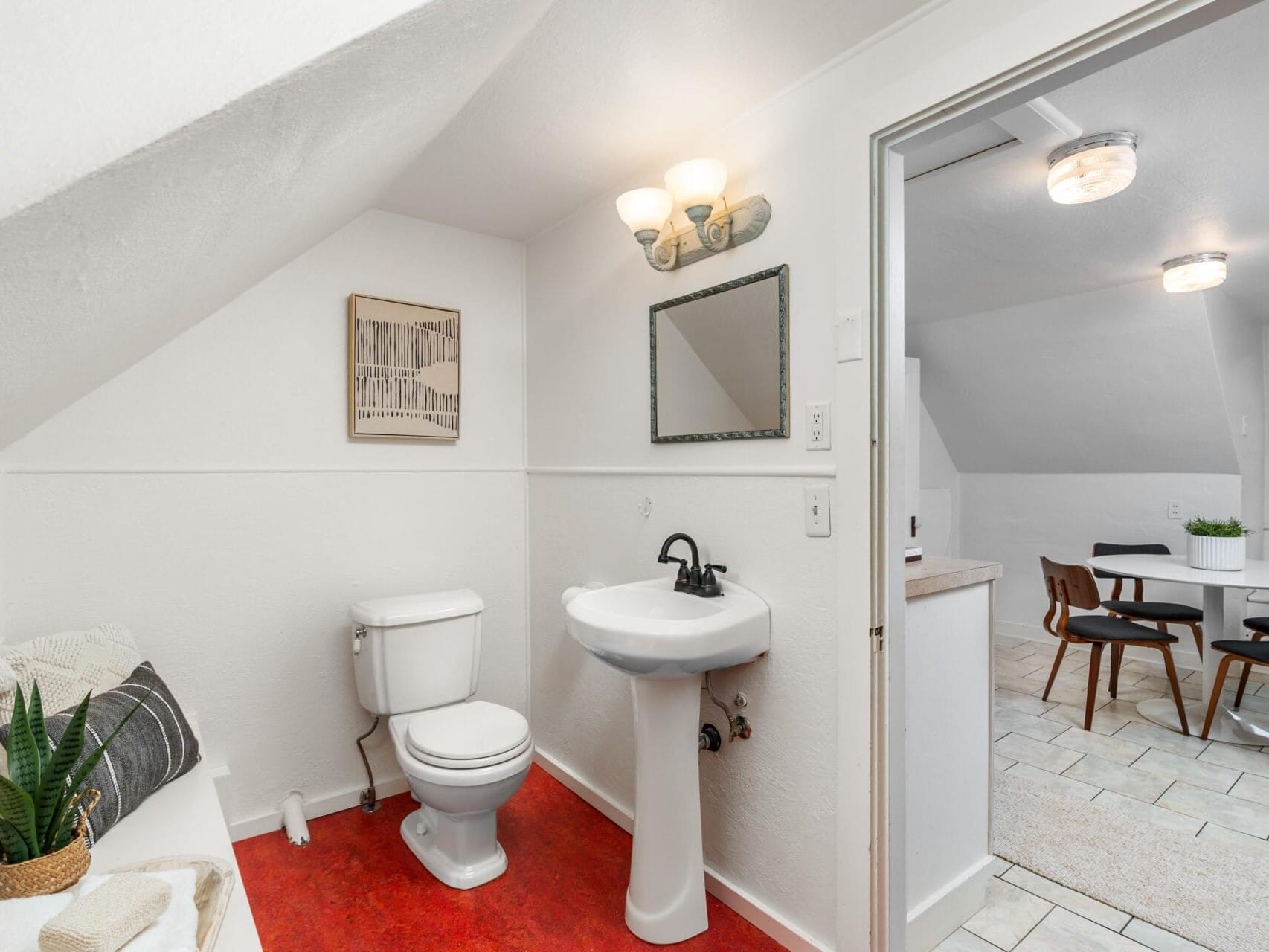 A small white bathroom with a toilet, pedestal sink, and black-framed mirror. Theres a red floor and a wall sconce above the mirror. A towel and potted plant sit on a bench. The room leads into a dining area with a table and chairs.