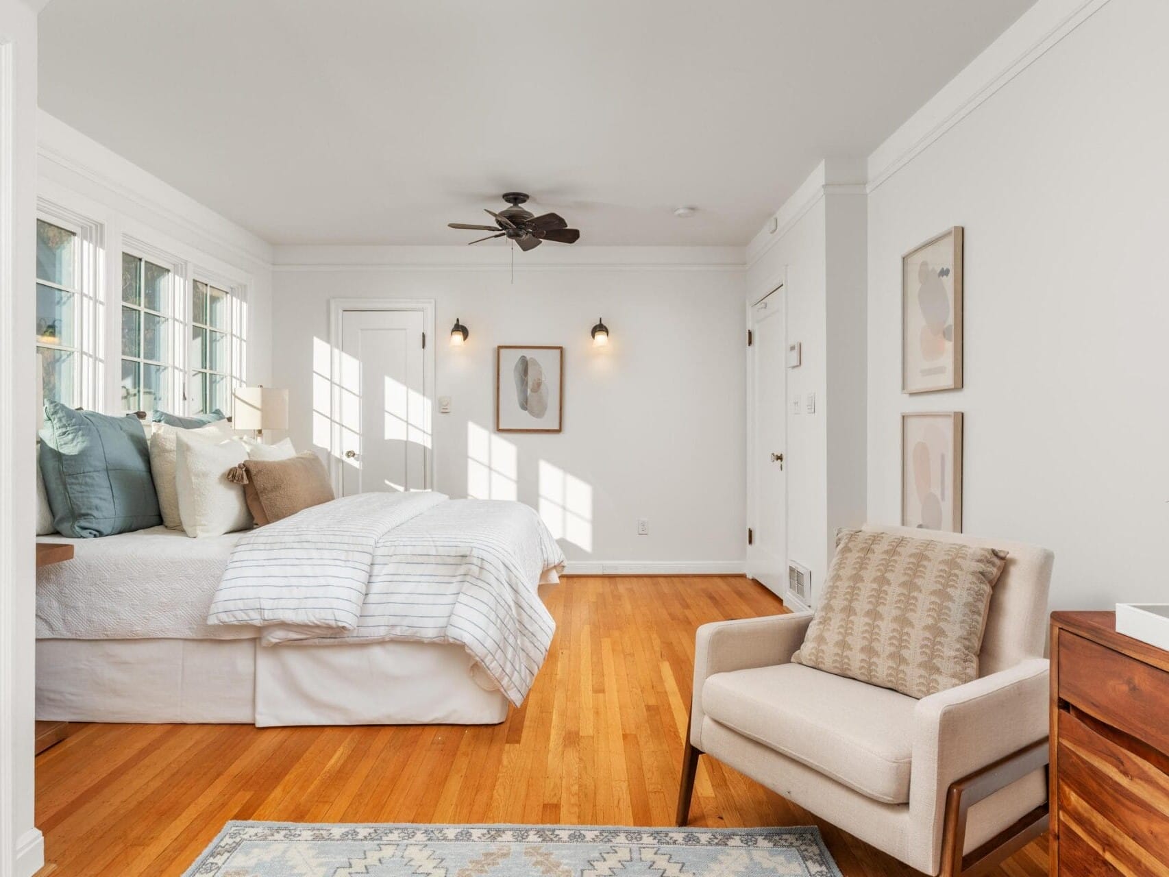 A bright bedroom with large windows, a cozy bed with striped bedding, and a white armchair on a wooden floor. Wall art and a ceiling fan enhance the minimalist decor. A small side table with a plant completes the space.