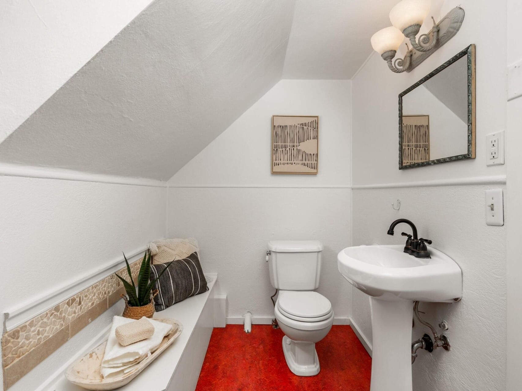 A small bathroom with white walls and ceiling. Features include a red floor, a white toilet, and a white pedestal sink with a black faucet. A mirror and light fixture are above the sink, and a decorative tray with a plant is on a ledge.