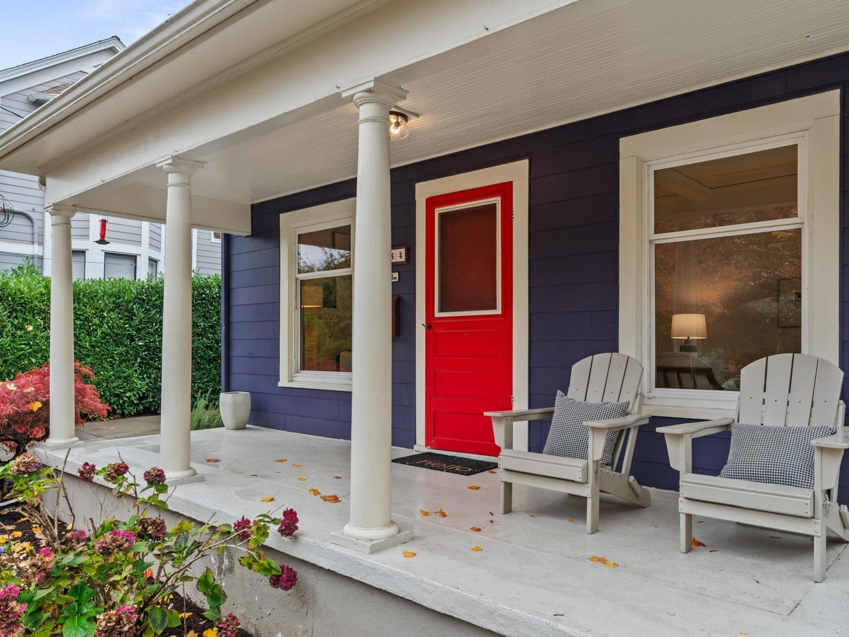 A cozy porch with a bright red door on a house with dark blue siding. Two white Adirondack chairs are set on the porch. The garden in front has red and purple flowers, and a neighboring house is visible on the left.