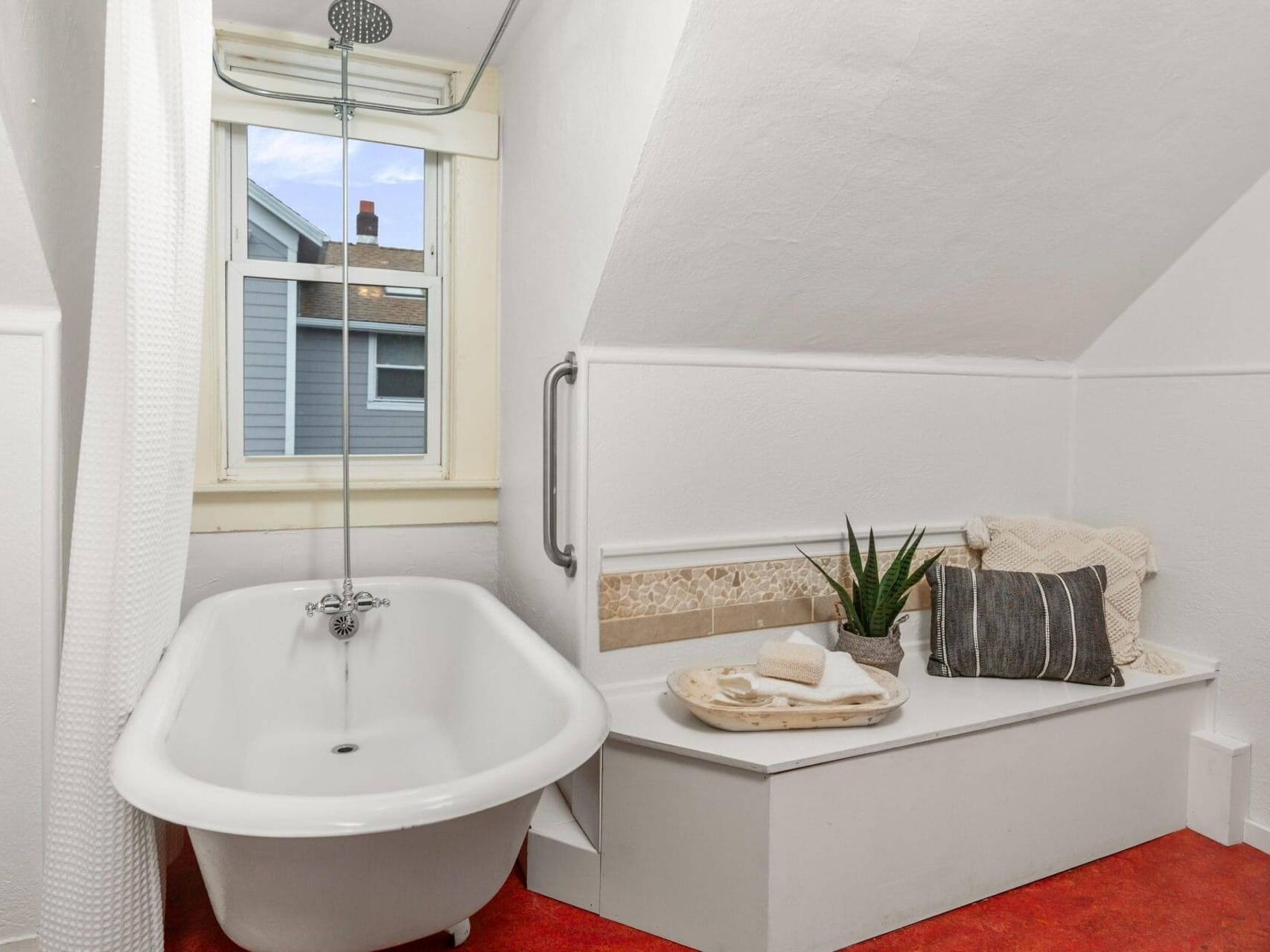 A cozy bathroom with a white clawfoot bathtub beneath a window. The room features a small seating area with pillows and decorative items, including a potted plant. The floor is covered with a red carpet.