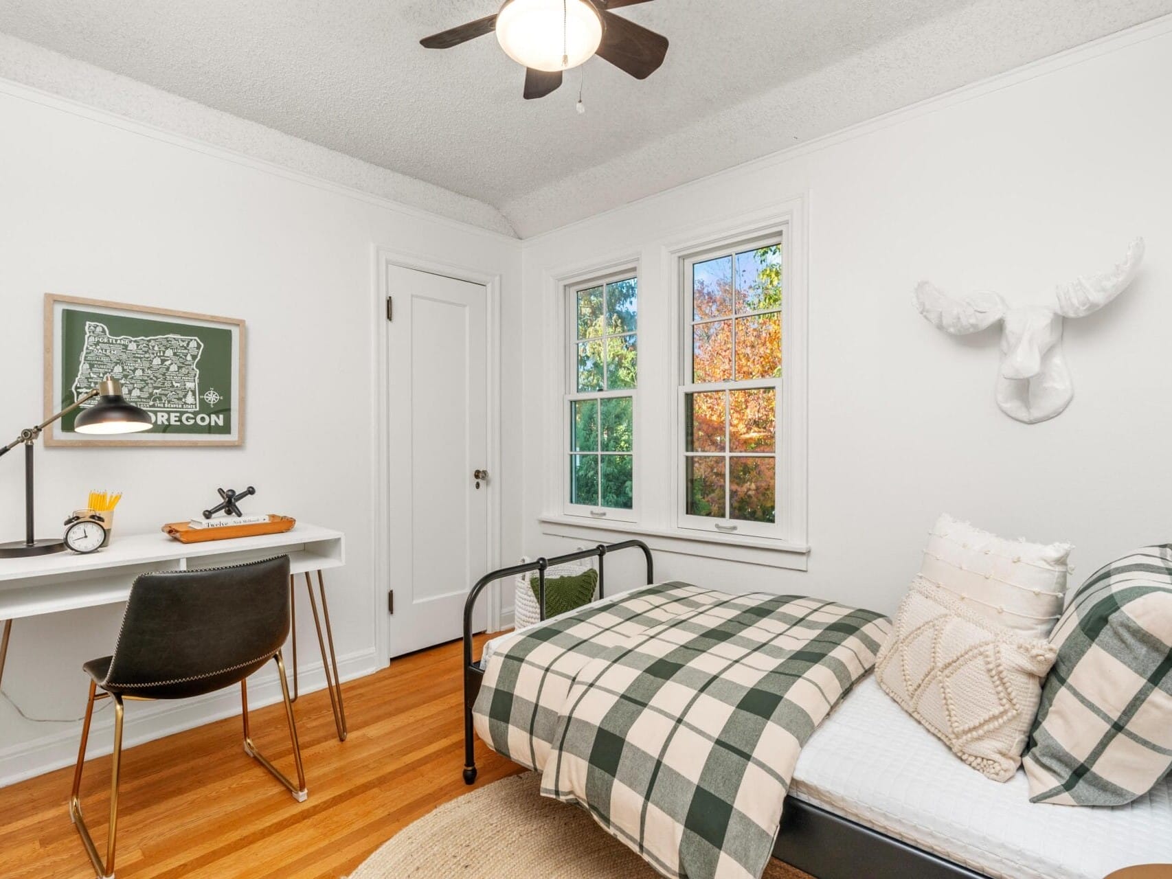 A cozy bedroom with wooden floors features a single bed with plaid bedding, a white desk with a lamp, and a black chair. A wall-mounted Oregon print and a white deer head sculpture add decor. Sunlight filters through two windows, illuminating the space.