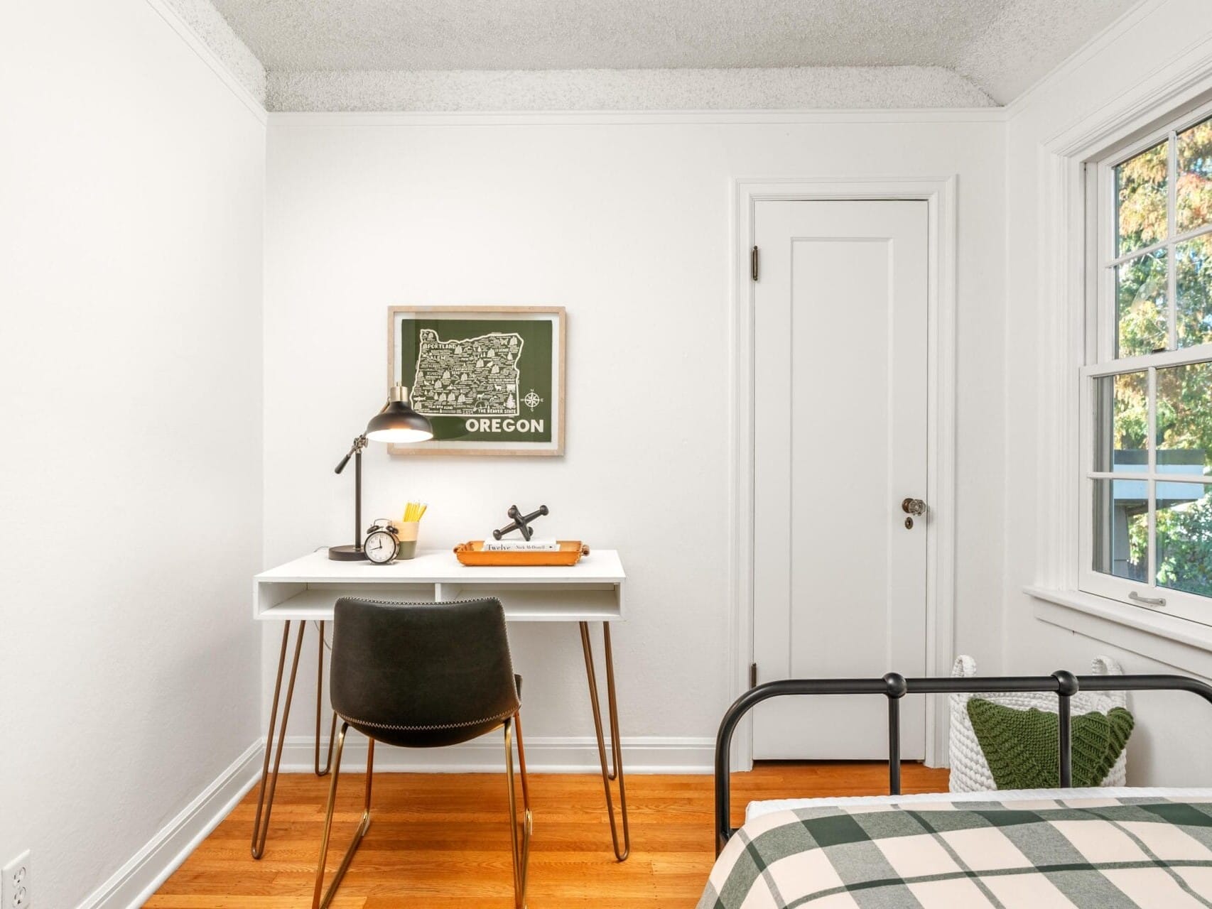 A small, cozy bedroom features a minimalistic workspace with a white desk, black chair, desk lamp, and decorations. A Oregon map hangs on the wall. The room has a bed with plaid bedding, wooden floor, and a window letting in natural light.