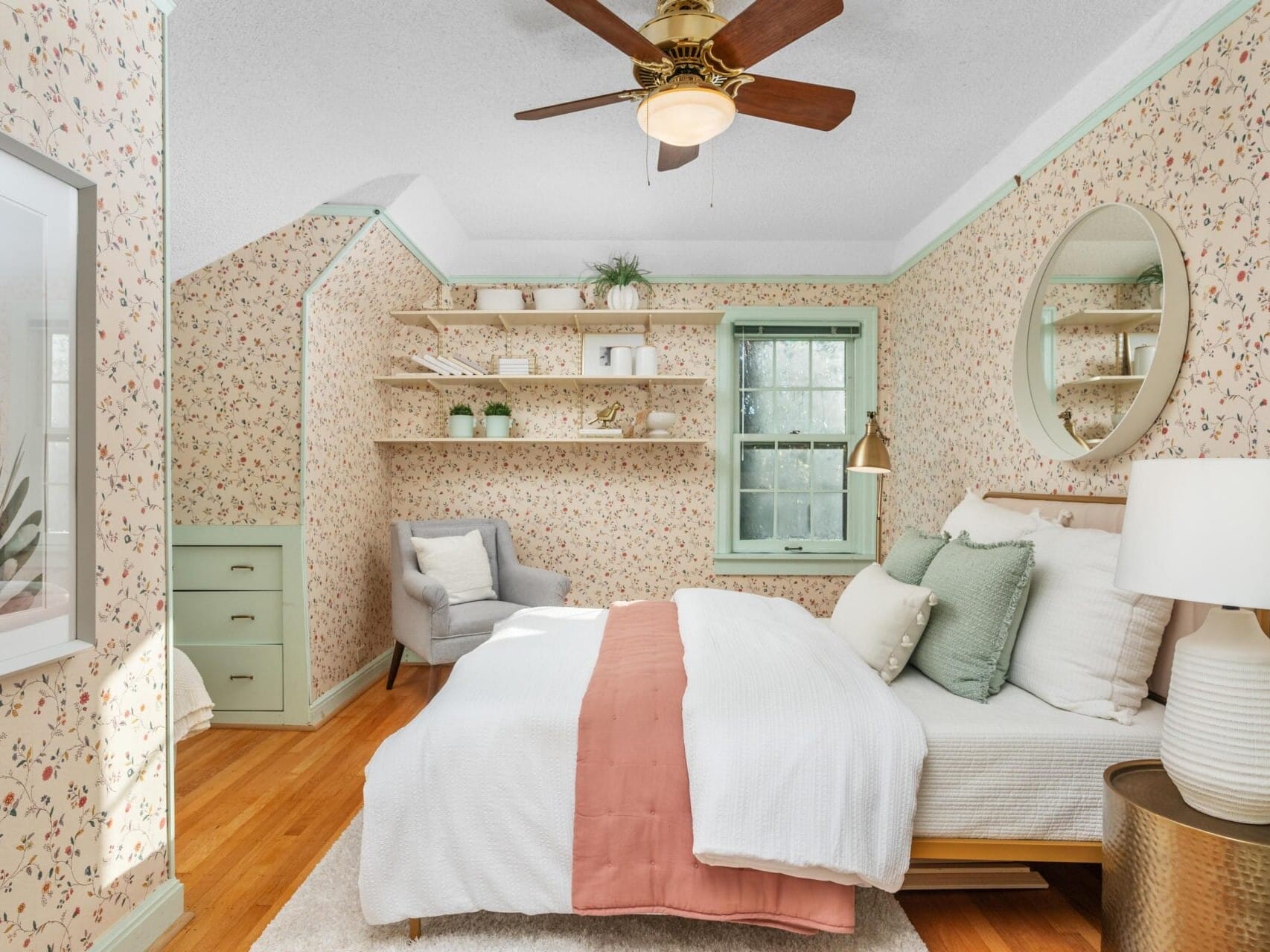 A cozy bedroom with floral wallpaper features a bed with white and coral bedding. There is a round mirror above the bed, a wooden ceiling fan, a corner chair, and wall shelves with decor. Light streams in through a window with green trim.