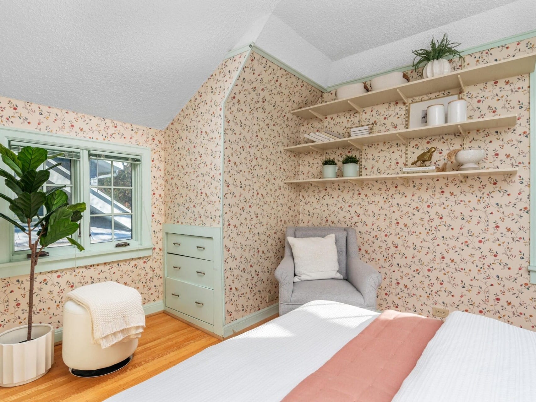 A cozy bedroom with floral wallpaper, a grey armchair, and a mint green chest of drawers. Open shelves hold plants and decor. A bed with white and peach bedding is partially visible. A potted tree sits beside a window with mint trim.