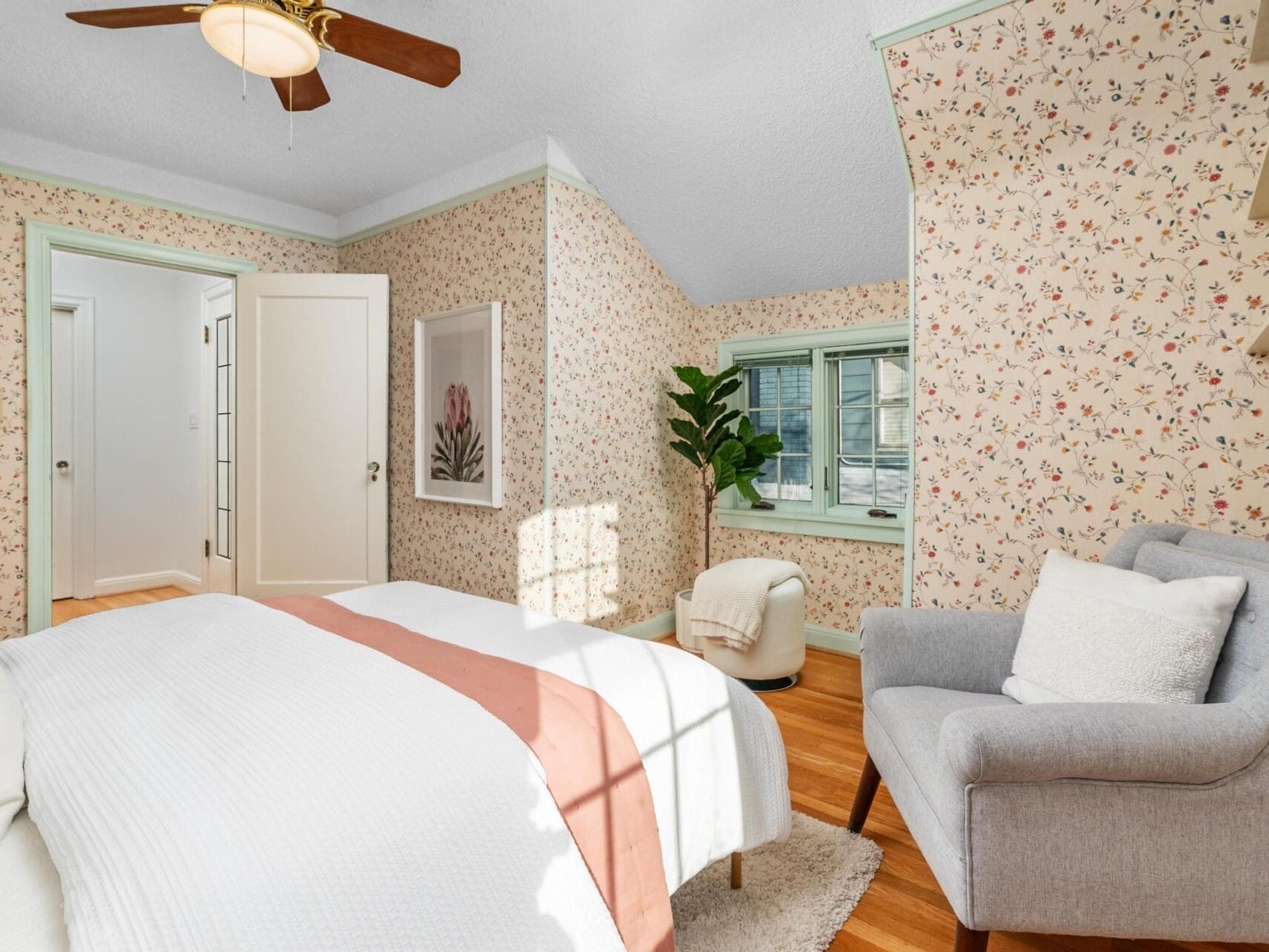 A cozy bedroom with floral wallpaper, a bed with white and peach bedding, a gray armchair, and a potted plant. A ceiling fan is above, and natural light streams through a window with light curtains. Doorway leads to another room.