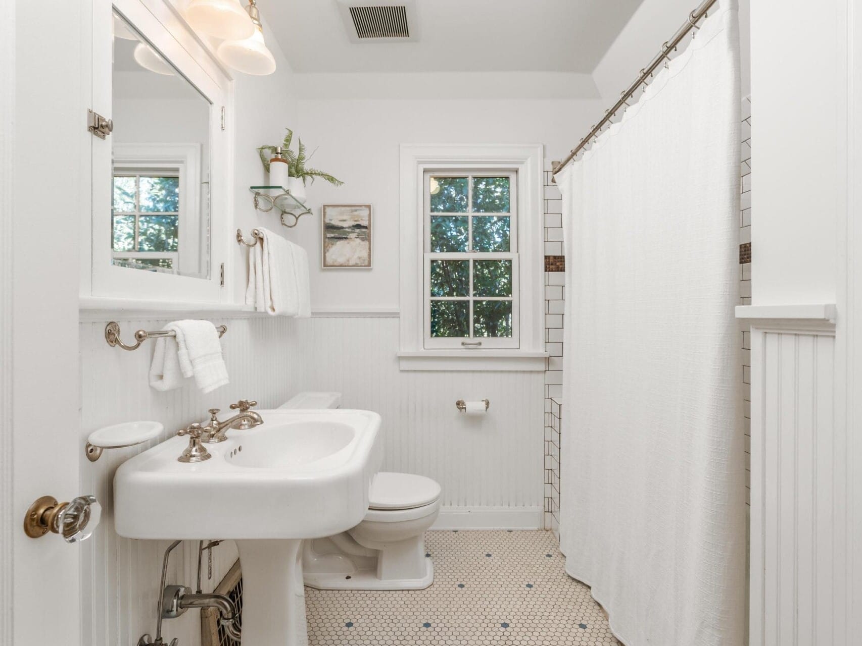 A bright white bathroom with a vintage-style sink and toilet. The floor is tiled with small hexagonal tiles. Theres a shower with a white curtain on the right and a window above the toilet. A small plant and towel hang on the wall.