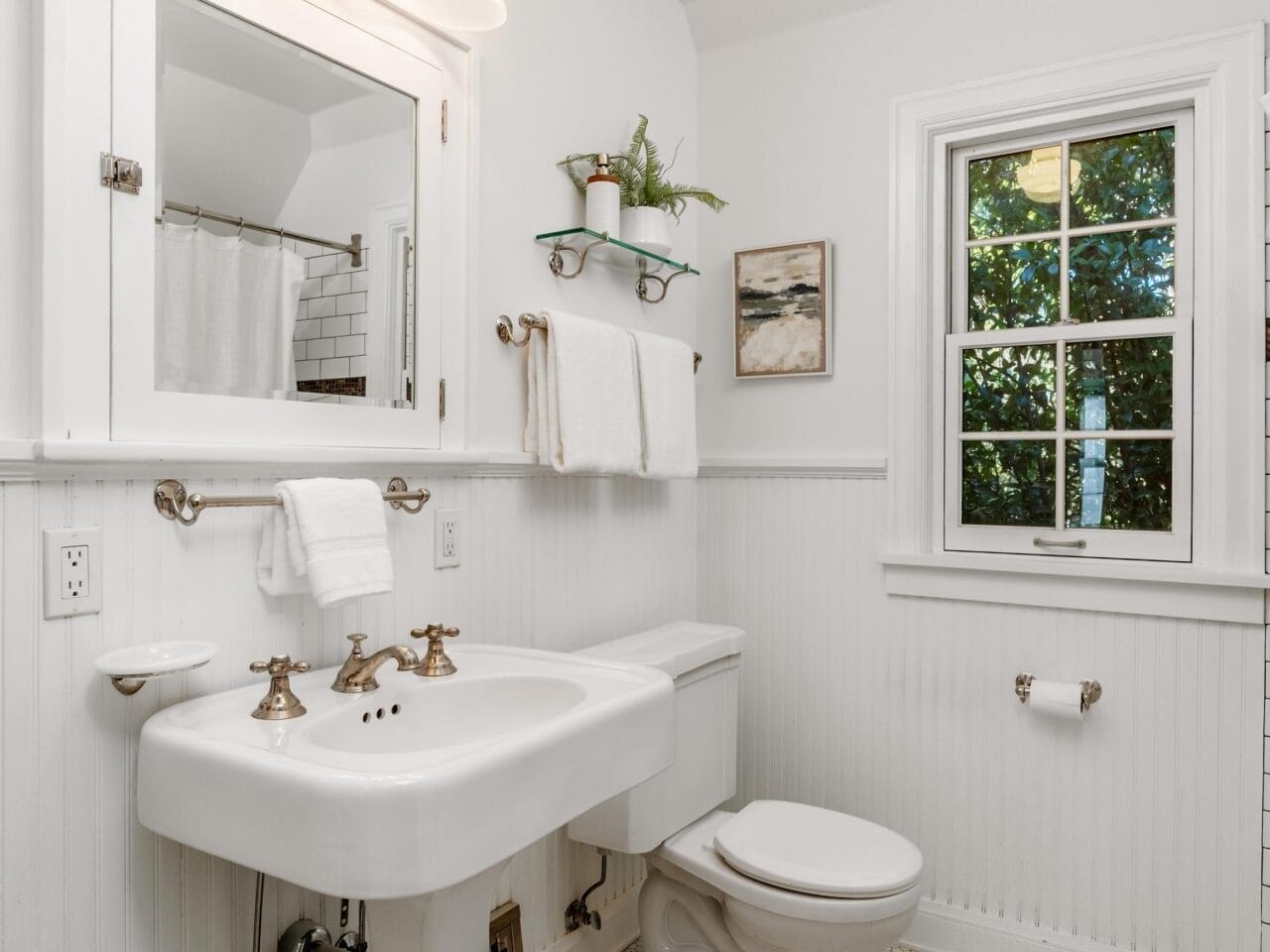 White bathroom with pedestal sink, toilet, and hexagonal tile floor. A mirror with lights is above the sink. Towel rack with towels, a small painting, and a window with a view of greenery are visible. Shelves hold plants and accessories.