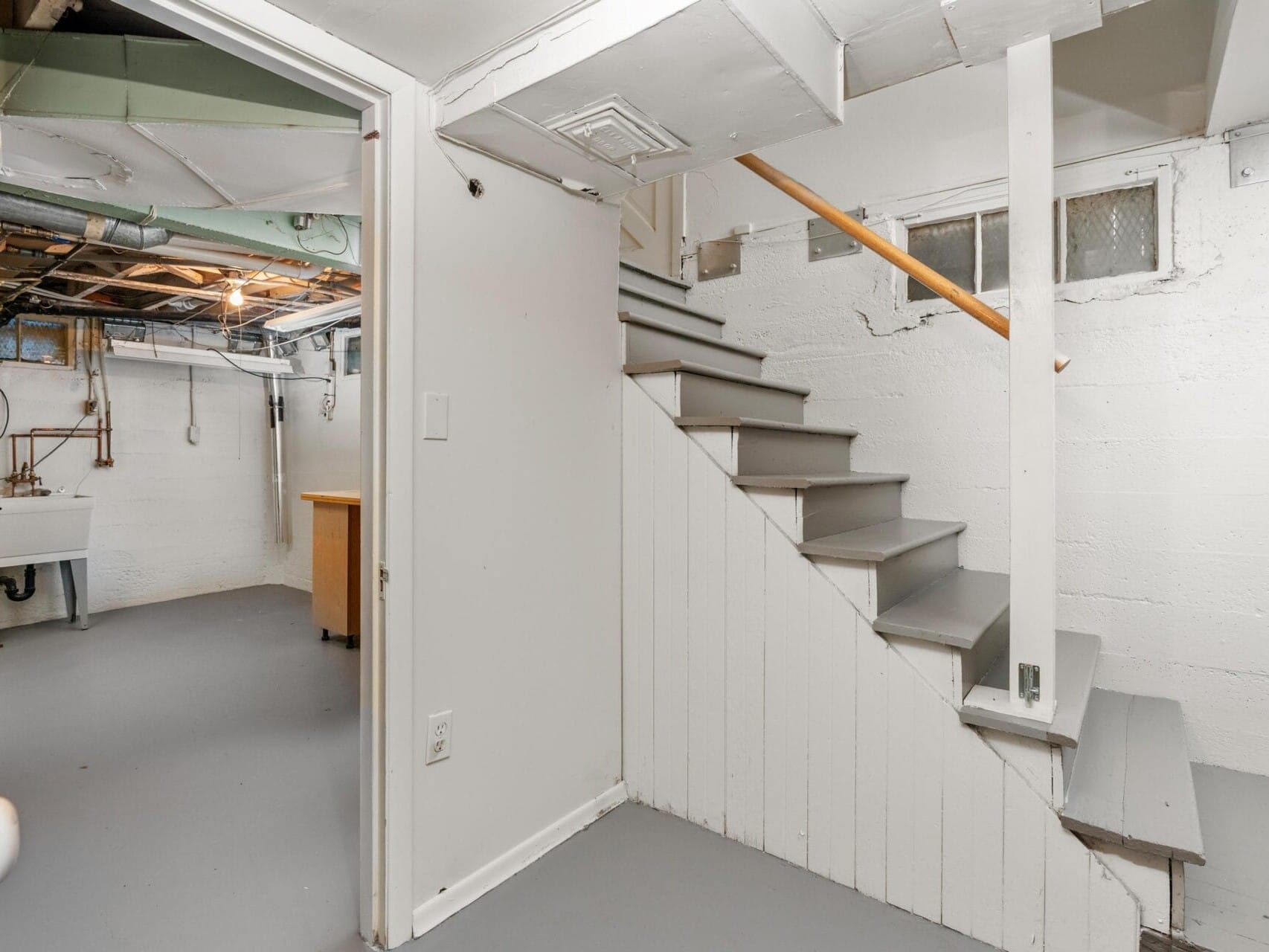 Basement room with white walls and gray floor, featuring a wooden staircase leading upwards. The room has exposed pipes and wires on the ceiling, a utility sink, and a small window. The lighting is dim, and a wooden cabinet is visible in the background.