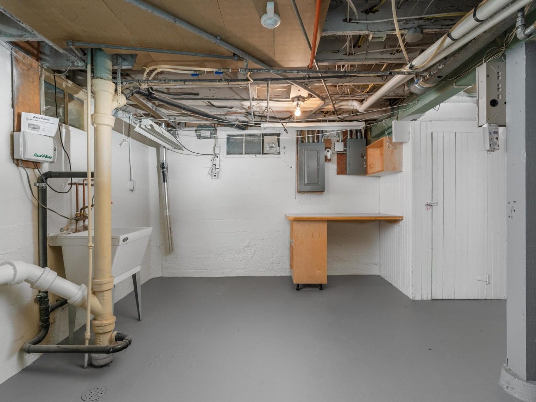 A basement with exposed pipes and wiring on the ceiling, a utility sink, a small wooden workbench, and a white door. The floor is painted gray, and the walls are light grey. A light bulb hangs from the ceiling providing illumination.