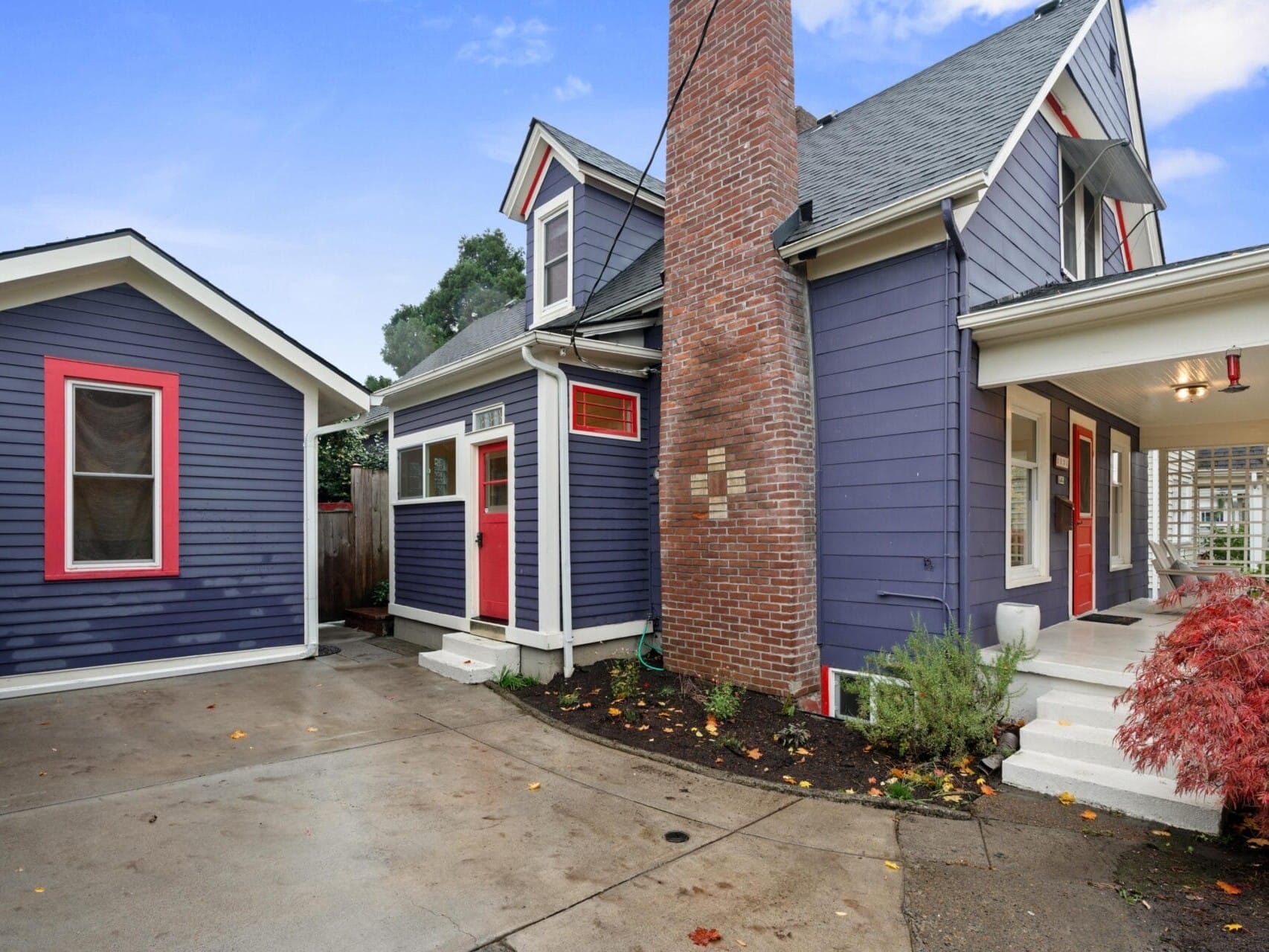 A charming blue house with red-trimmed windows and a central brick chimney. The house features a covered porch, a detached garage, white stairs, and a small red tree in the front yard. A concrete driveway leads to the entrance.