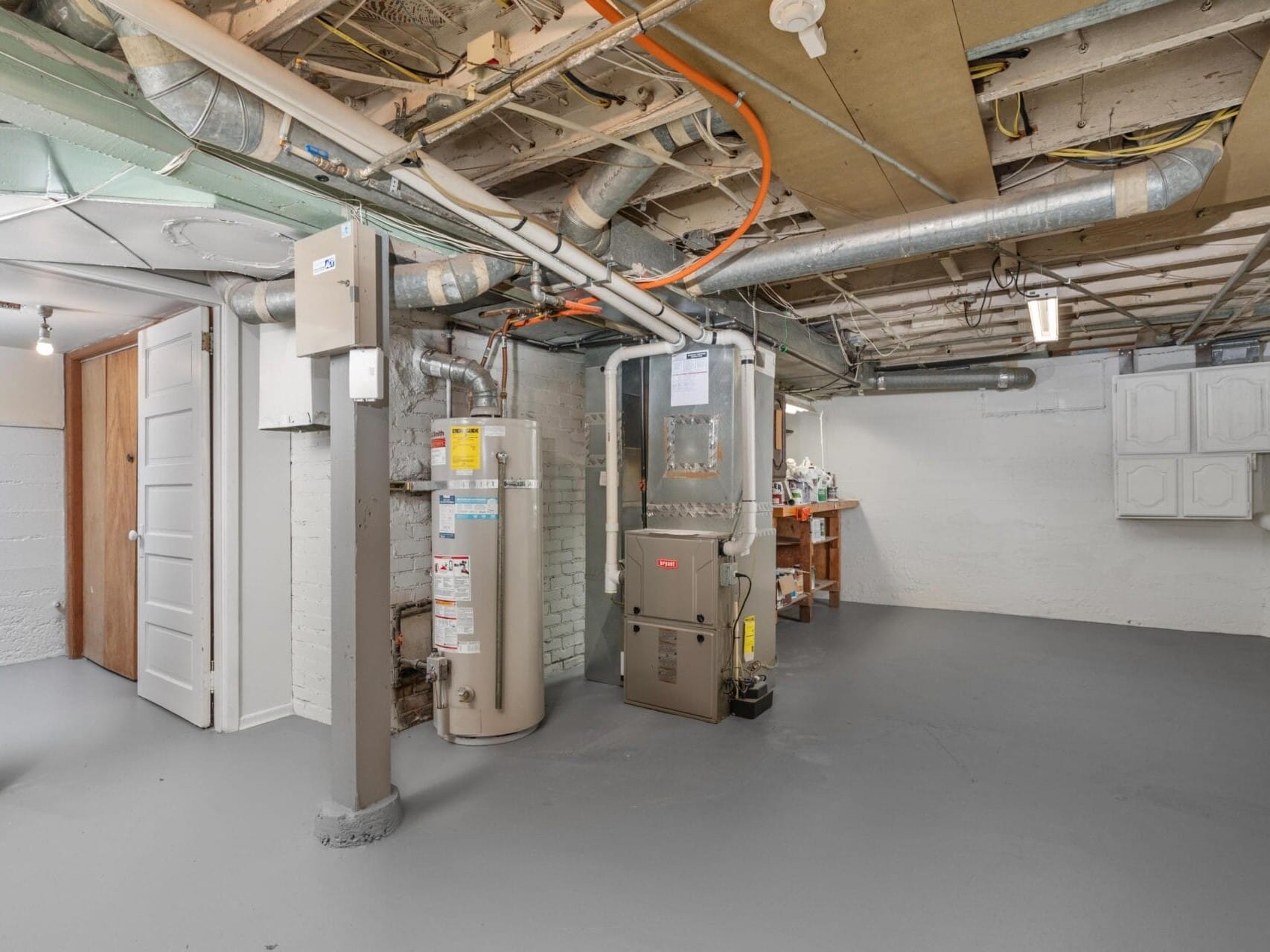 A clean, unfinished basement with exposed pipes and wiring. There is a water heater and furnace in the center, with a workbench and tools in the background. The floor is painted gray, and a partially open door is visible on the left.