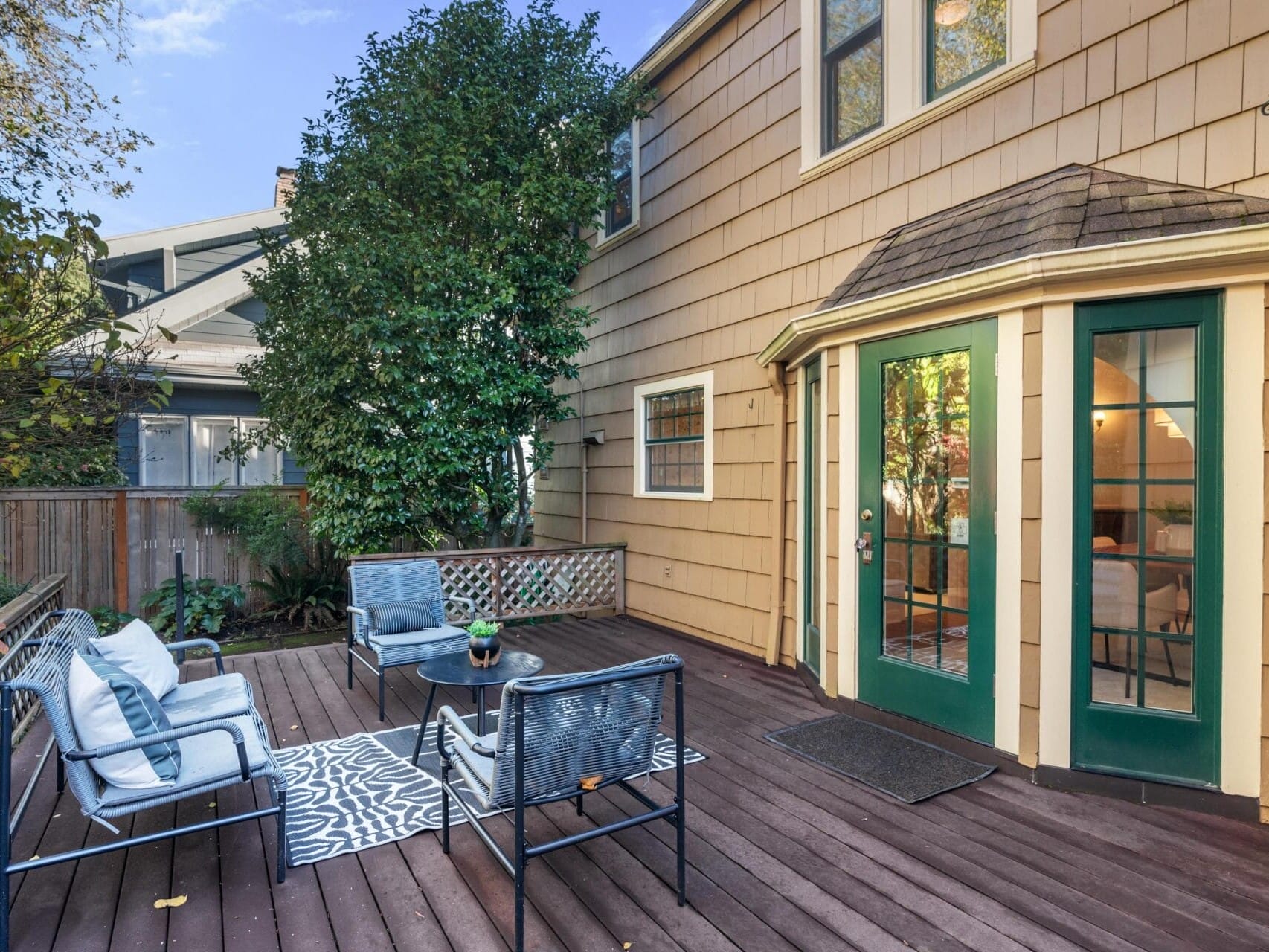 A cozy backyard deck featuring a seating area with blue cushioned chairs around a small table on a patterned rug. The deck is attached to a beige house with green-framed double doors and surrounded by a wooden fence and lush greenery.