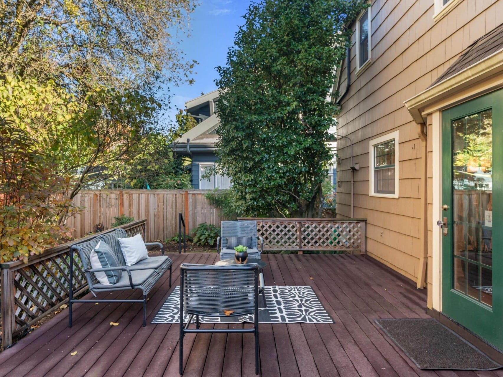 A wooden deck in a backyard with outdoor furniture, including a sofa, chairs, and a small table on a patterned rug. The deck is surrounded by greenery and a wooden fence, next to a house with tan siding and a green door.