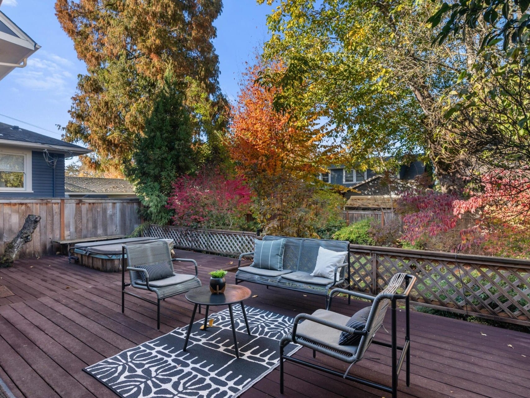 Outdoor patio with wooden decking, featuring a seating area with a small table, two chairs, and a loveseat. A patterned rug is beneath the furniture. Lush autumn trees with vibrant leaves surround the area, creating a cozy, inviting atmosphere.