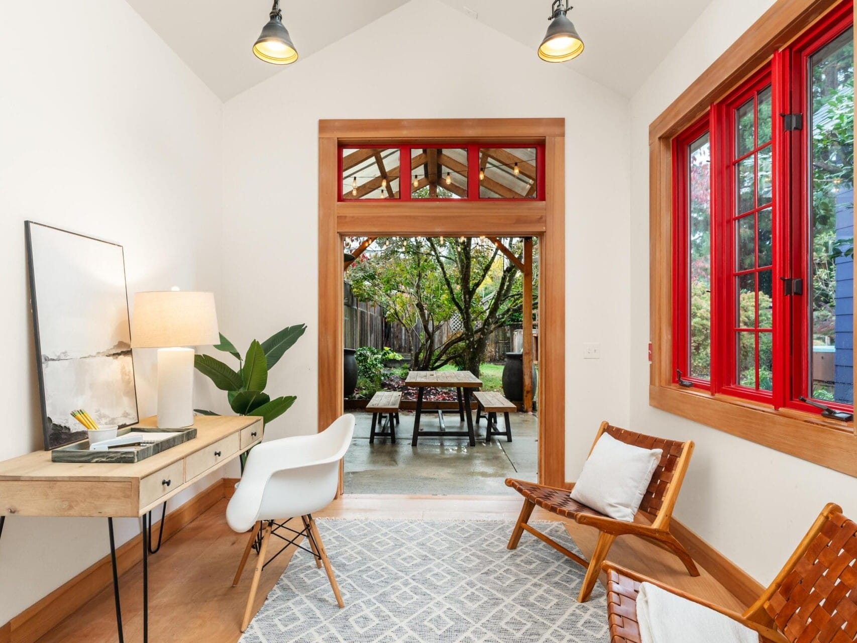 Bright sunroom with a wooden desk, chair, lamp, and art piece. Floor-to-ceiling windows with red frames open to a garden patio with outdoor seating. Two wooden lounge chairs and a patterned rug complete the cozy, natural setting.