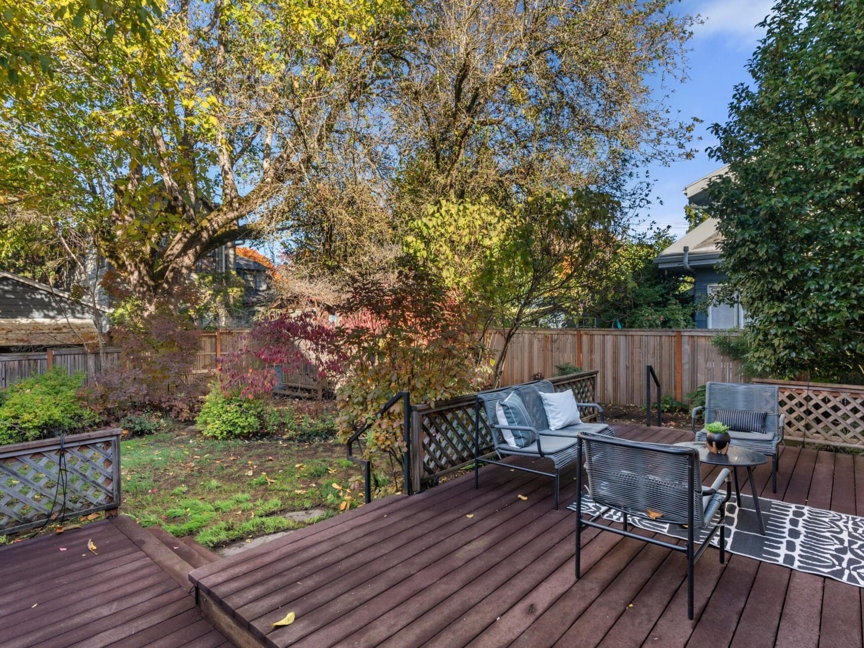 A wooden deck with outdoor seating, including a table and chairs, overlooks a backyard with lush trees and a wooden fence. The area is adorned with a patterned rug and has a peaceful, natural ambiance.