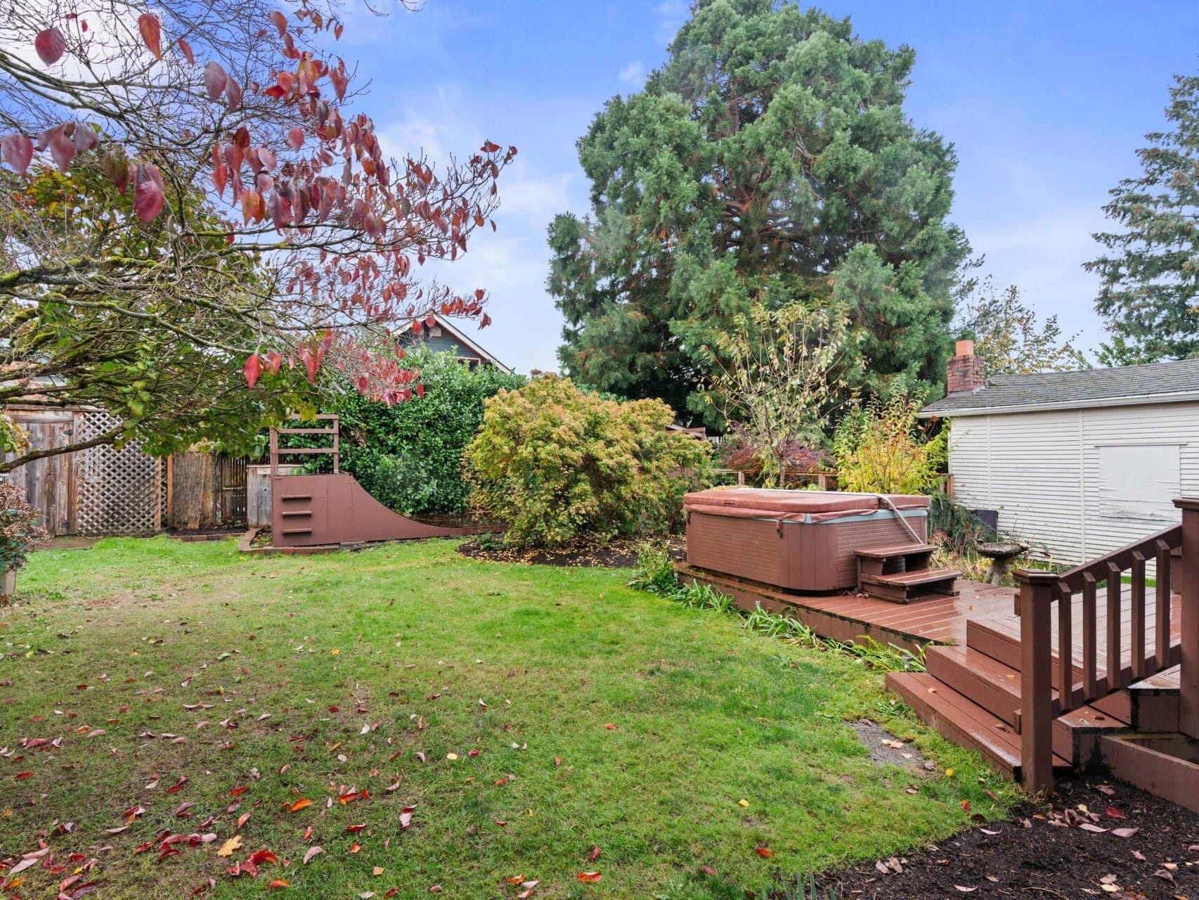 A backyard with green grass, trees with autumn leaves, a wooden play structure, and a wooden deck featuring a hot tub. A large evergreen tree and a small garden are visible, along with a white shed or building on the right.