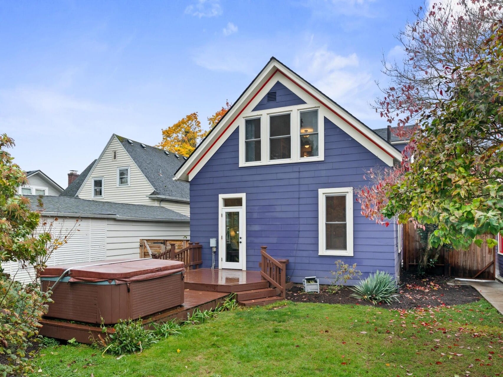 A blue two-story house with white trim sits in a green yard. A wooden deck leads to a hot tub on the left. Trees with autumn leaves surround the property under a blue sky.