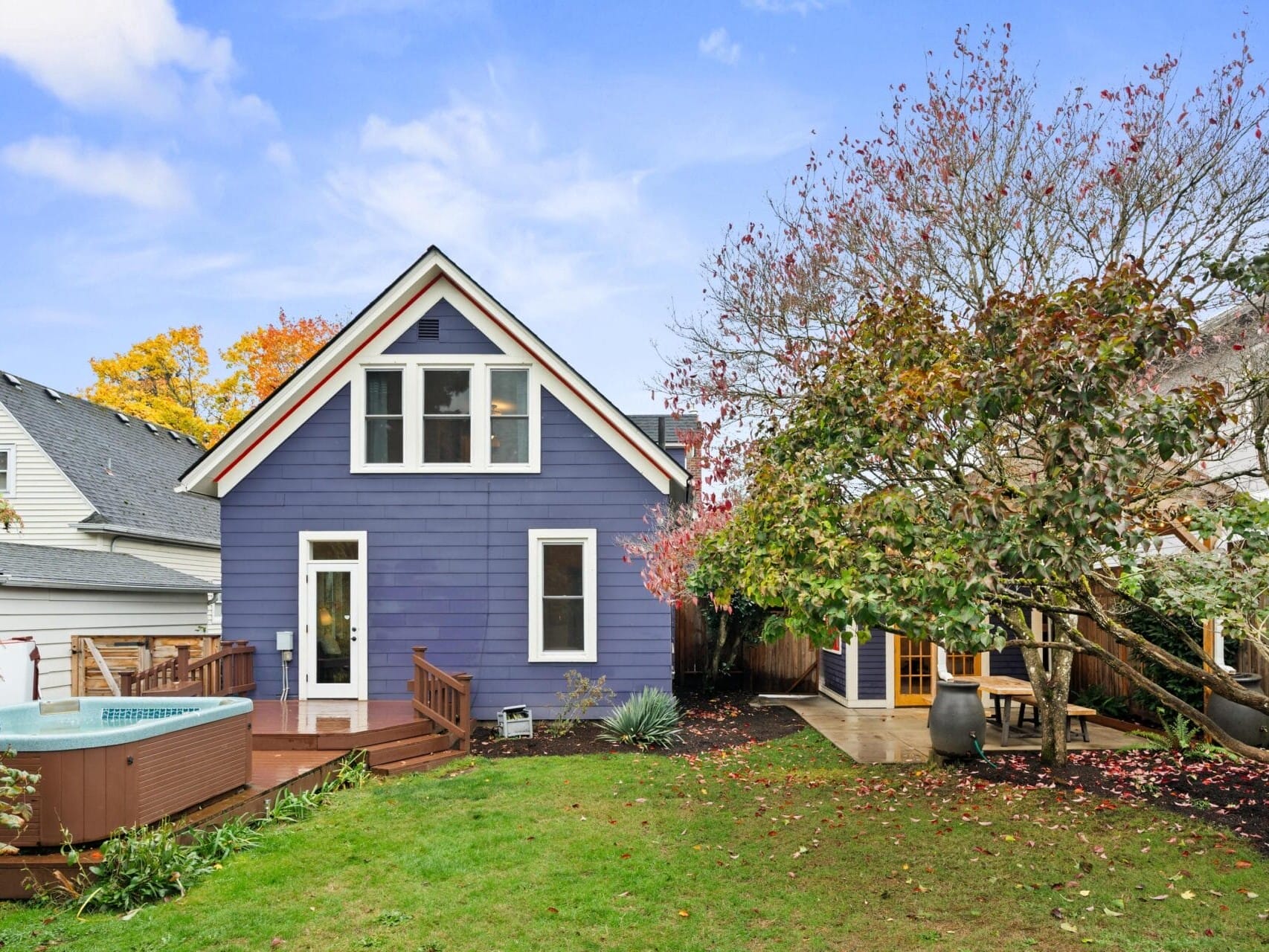 A charming purple house with white trim sits in a grassy backyard area. Next to the house is a hot tub with steps leading up to it. Trees with autumnal leaves surround the property under a clear blue sky.