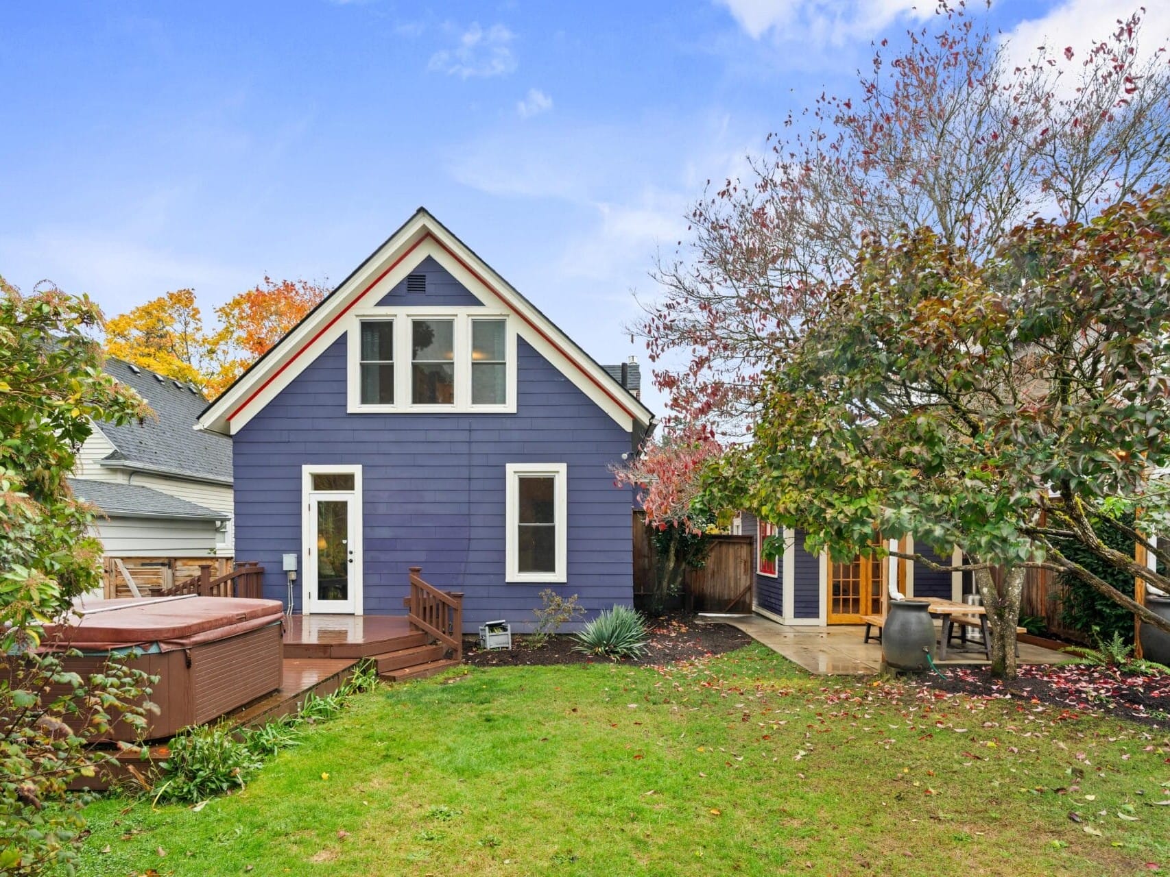 A house with blue siding, white trim, and a steep roof sits in a yard with green grass. Theres a wooden deck with a hot tub and a tree with autumn foliage nearby. The sky is partly cloudy.