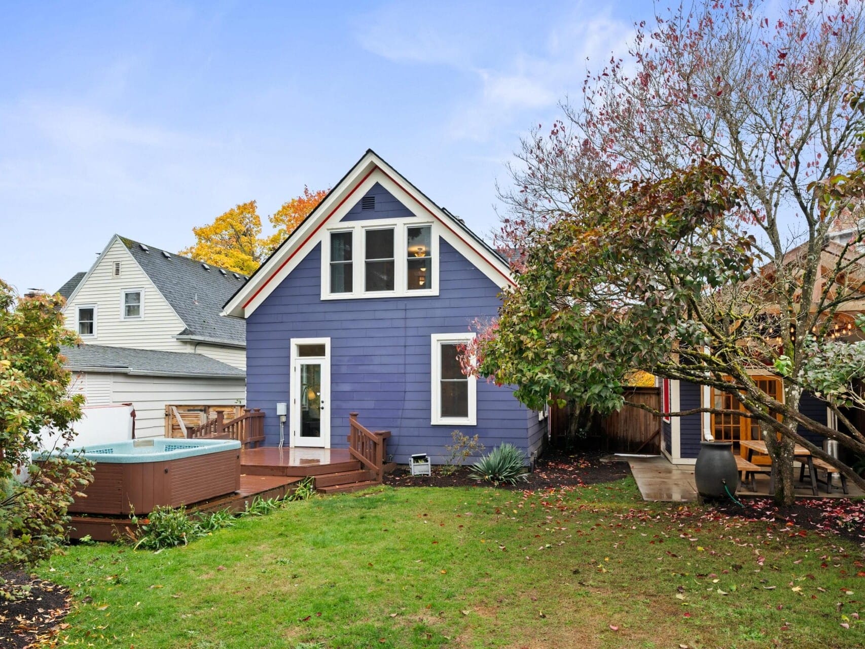 A charming blue house with a triangular roof and white trim is surrounded by lush trees. Theres a wooden deck at the back with a small pool. The yard is green, with some fallen leaves, under a partly cloudy sky.